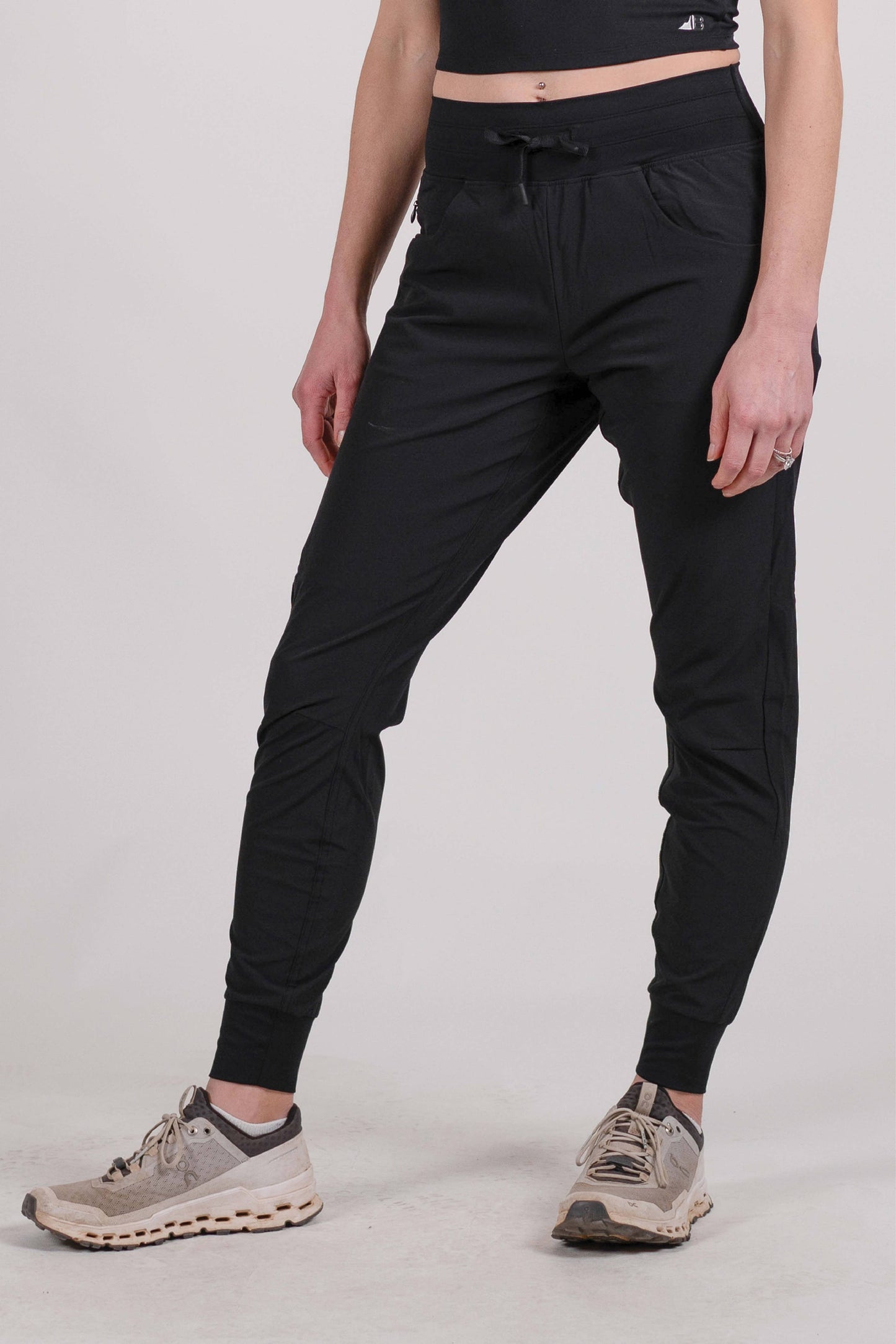 A person wearing Belong Designs' Rocky Mountain Joggers, crafted from durable and stretchy material, stands against a plain background. They are dressed in a dark green cropped top paired with matching athletic pants featuring a drawstring, complemented by brown hiking boots with teal laces. The image is cropped to show the midriff down.
