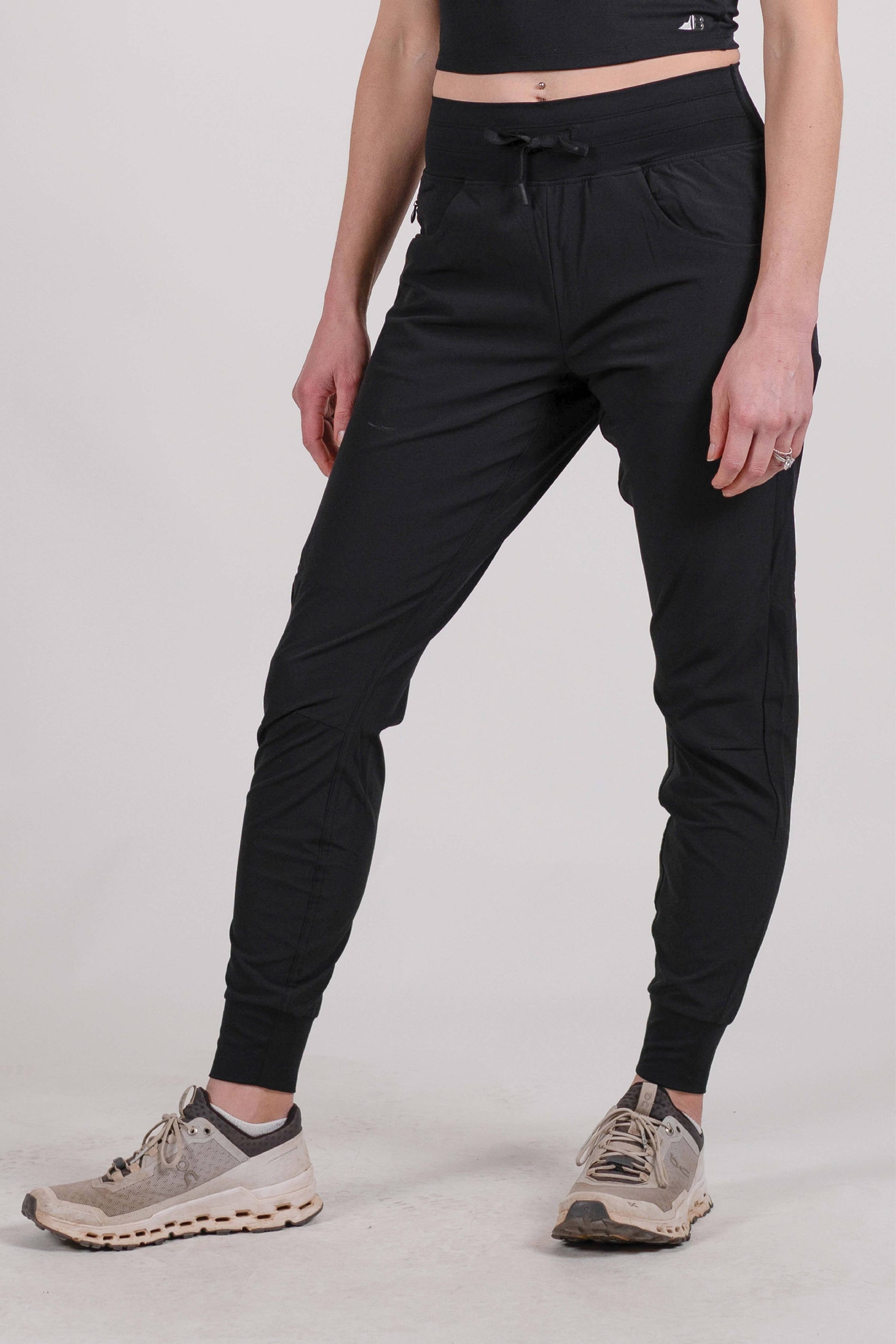 A person wearing Belong Designs' Rocky Mountain Joggers, crafted from durable and stretchy material, stands against a plain background. They are dressed in a dark green cropped top paired with matching athletic pants featuring a drawstring, complemented by brown hiking boots with teal laces. The image is cropped to show the midriff down.
