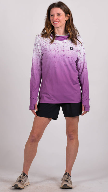 A person in a long-sleeve Sun Hoodie by Belong Designs, featuring a color-blocked design with white, maroon, and gray sections, a black logo on the chest, and wearing dark green shorts stands with hands in pockets. The background is plain white.