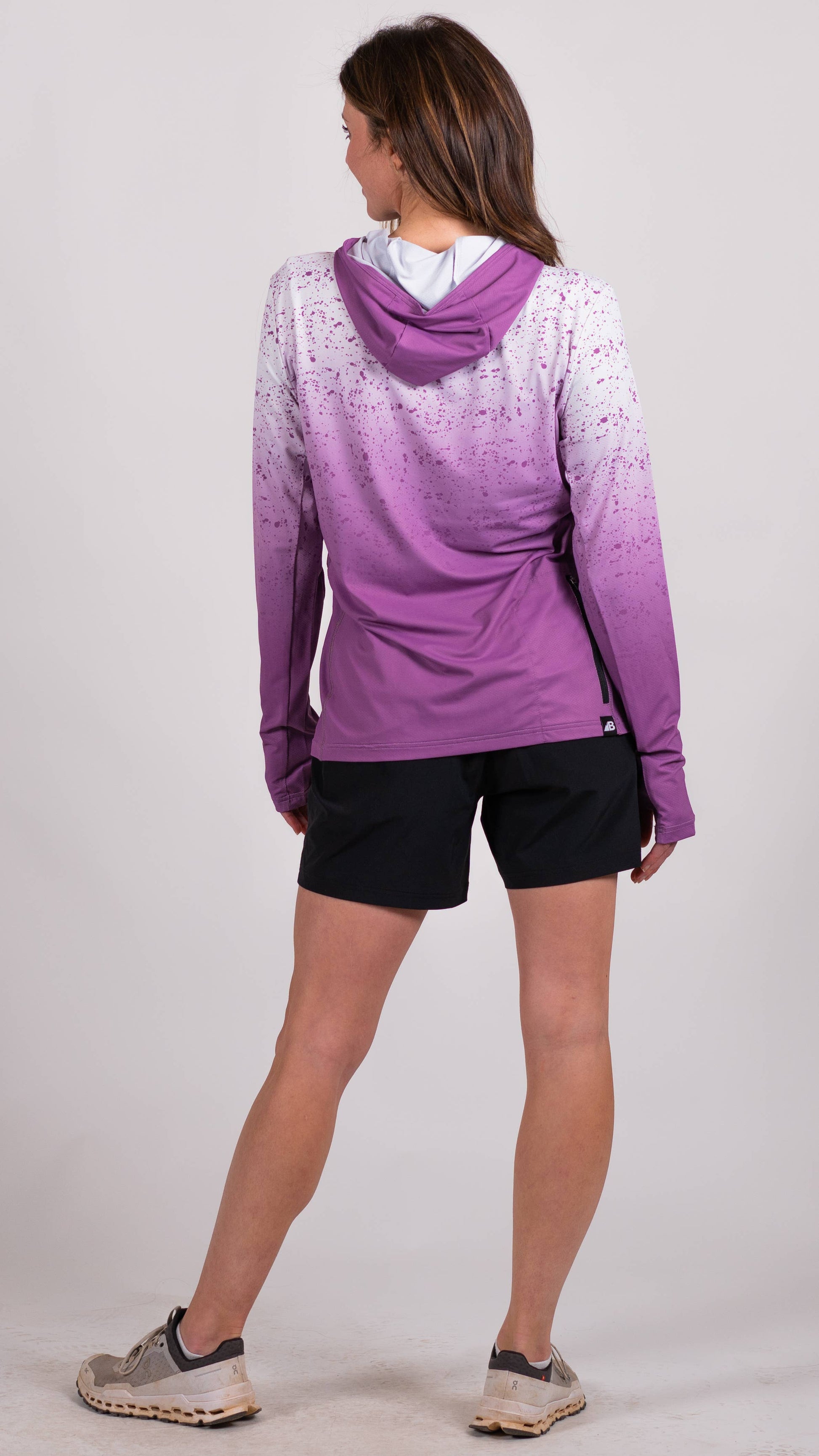 A person in a long-sleeve Sun Hoodie by Belong Designs, featuring a color-blocked design with white, maroon, and gray sections, a black logo on the chest, and wearing dark green shorts stands with hands in pockets. The background is plain white.