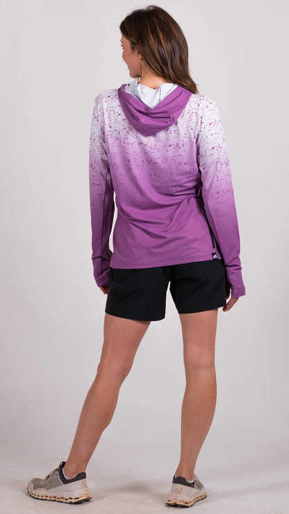 A person in a long-sleeve Sun Hoodie by Belong Designs, featuring a color-blocked design with white, maroon, and gray sections, a black logo on the chest, and wearing dark green shorts stands with hands in pockets. The background is plain white.