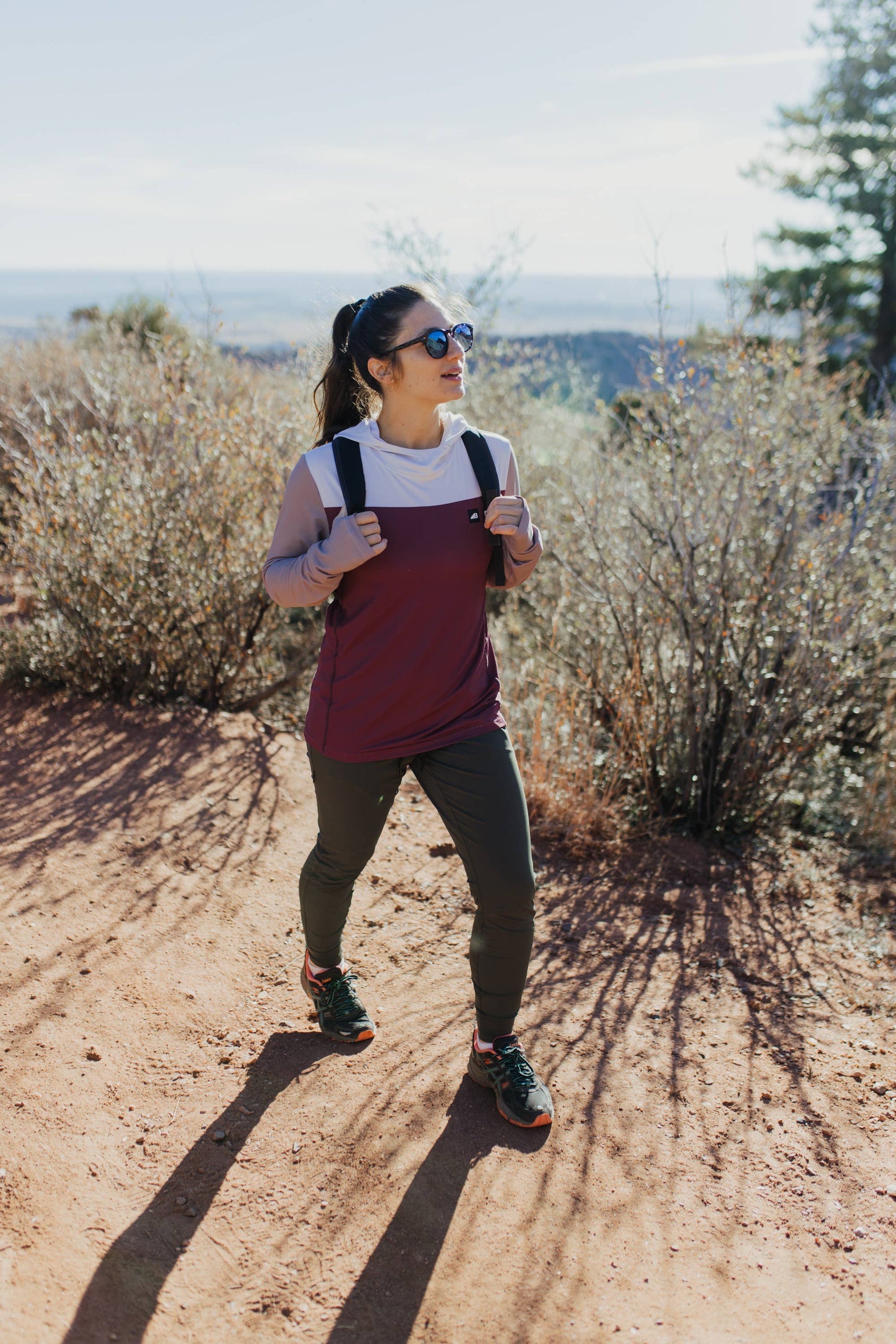 A person in a long-sleeve Sun Hoodie by Belong Designs, featuring a color-blocked design with white, maroon, and gray sections, a black logo on the chest, and wearing dark green shorts stands with hands in pockets. The background is plain white.