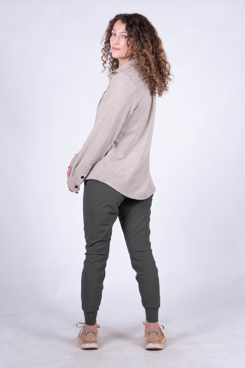 A person with curly hair stands against a plain background, smiling and looking at the camera. They are wearing the Sherman Fleece Button Up from Belong Designs, olive green pants, and beige shoes, with their hands in their pockets.
