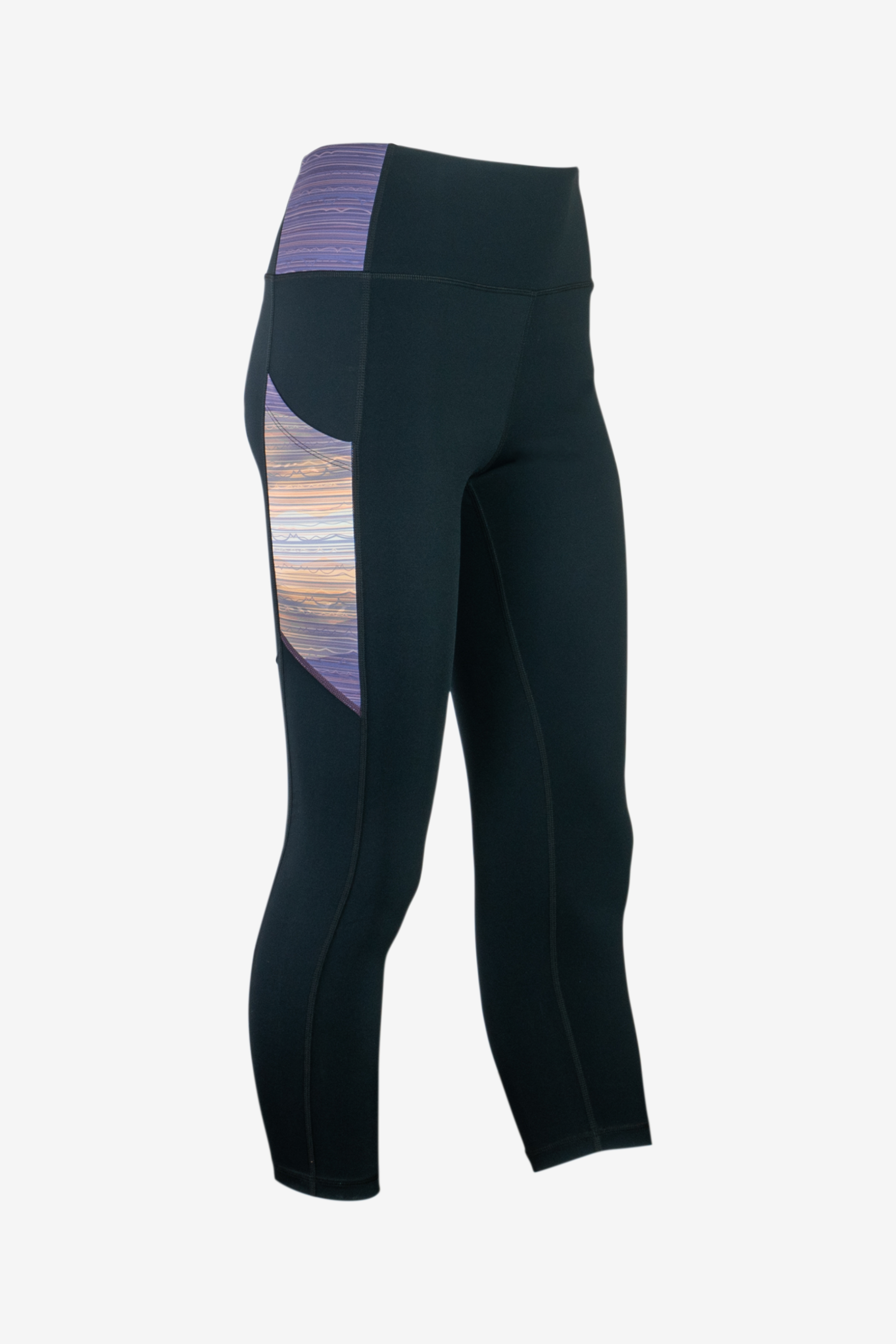 The Trailhead 7/8 Leggings from Belong Designs are black athletic leggings with a high waist and calf-length cut. They feature a colorful striped pattern in shades of purple and blue on the waist and leg panels. Crafted from PrimaFlex material, these multi-pocketed leggings also display a small logo on the left leg against a white background.