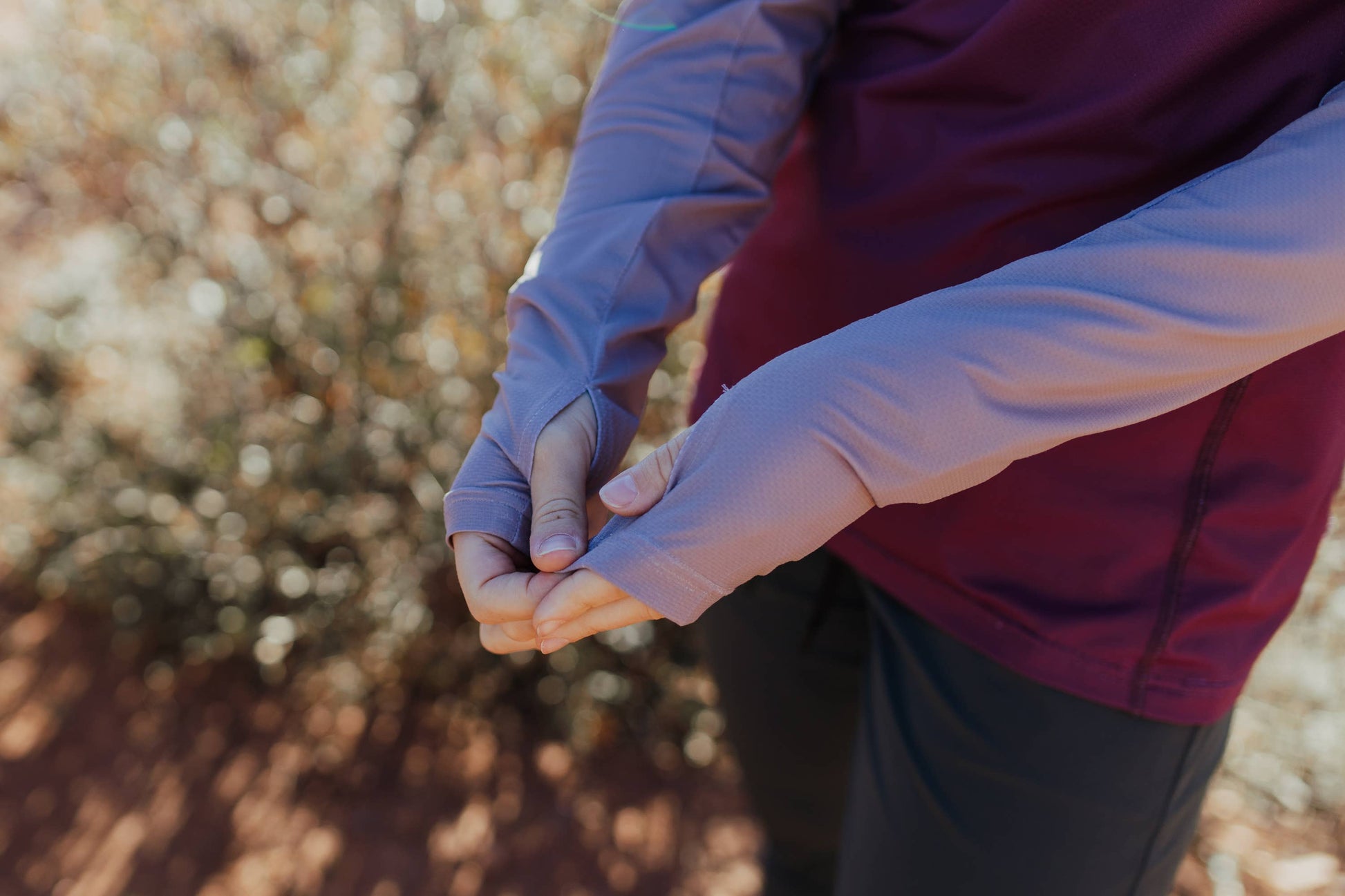 A person in a long-sleeve Sun Hoodie by Belong Designs, featuring a color-blocked design with white, maroon, and gray sections, a black logo on the chest, and wearing dark green shorts stands with hands in pockets. The background is plain white.
