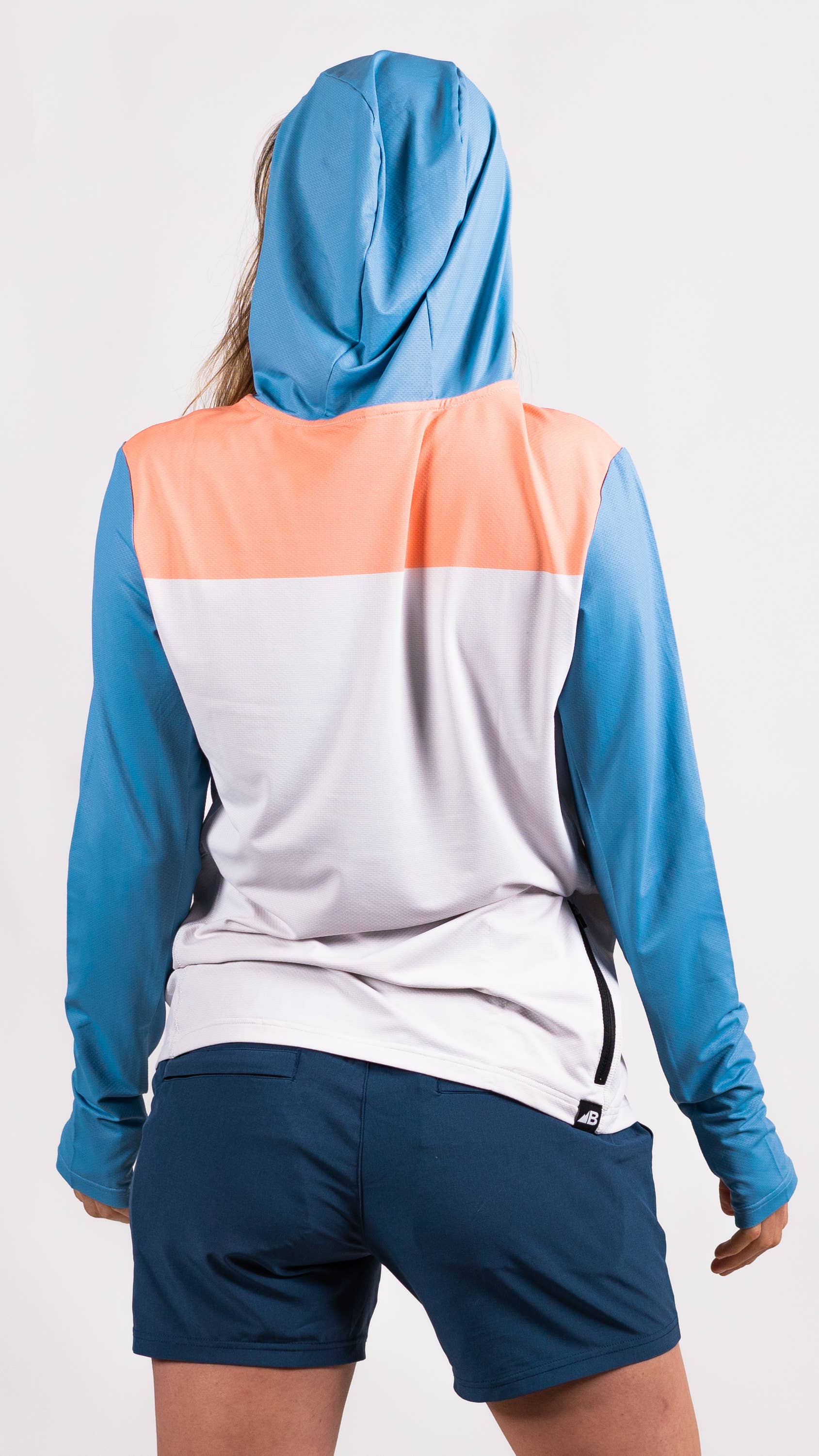 A person in a long-sleeve Sun Hoodie by Belong Designs, featuring a color-blocked design with white, maroon, and gray sections, a black logo on the chest, and wearing dark green shorts stands with hands in pockets. The background is plain white.