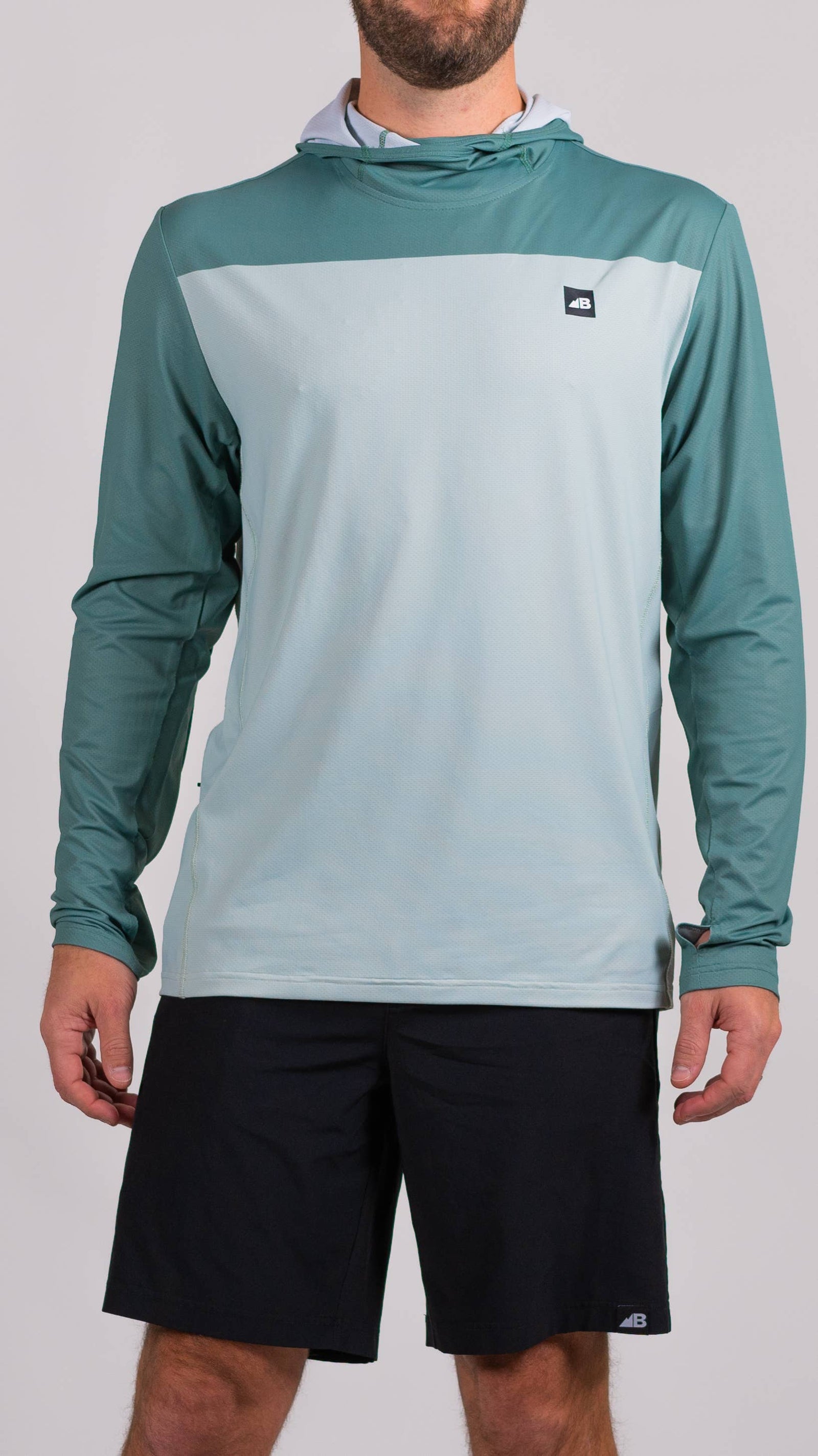 A man is wearing a Belong Designs Men's Sun Hoodie in Green Block, paired with black shorts. The hoodie features a small black logo on the left chest and offers UPF 50+ protection. He is standing against a plain, light-colored background, ready for outdoor activities. His head is not visible in the image.