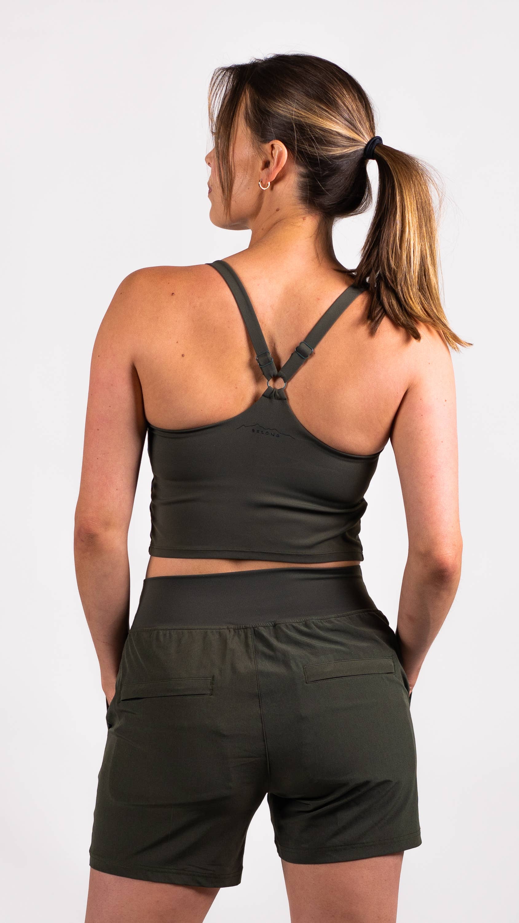 A person with long hair is dressed in a blue workout outfit from Belong Designs, consisting of the Trailhead Tek Tank in Steel Blue with adjustable straps and jogger-style pants. They are posing against a simple, white background, wearing sneakers. The person is looking to the side with one hand lightly resting near their neck.