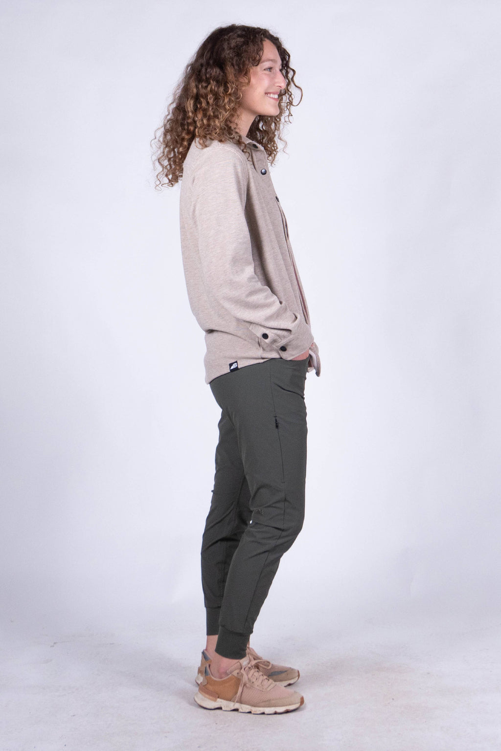 A person with curly hair stands against a plain background, smiling and looking at the camera. They are wearing the Sherman Fleece Button Up from Belong Designs, olive green pants, and beige shoes, with their hands in their pockets.