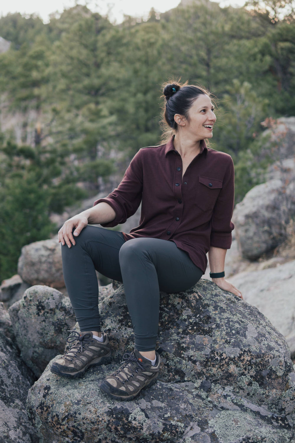 A person with curly hair stands against a plain background, smiling and looking at the camera. They are wearing the Sherman Fleece Button Up from Belong Designs, olive green pants, and beige shoes, with their hands in their pockets.