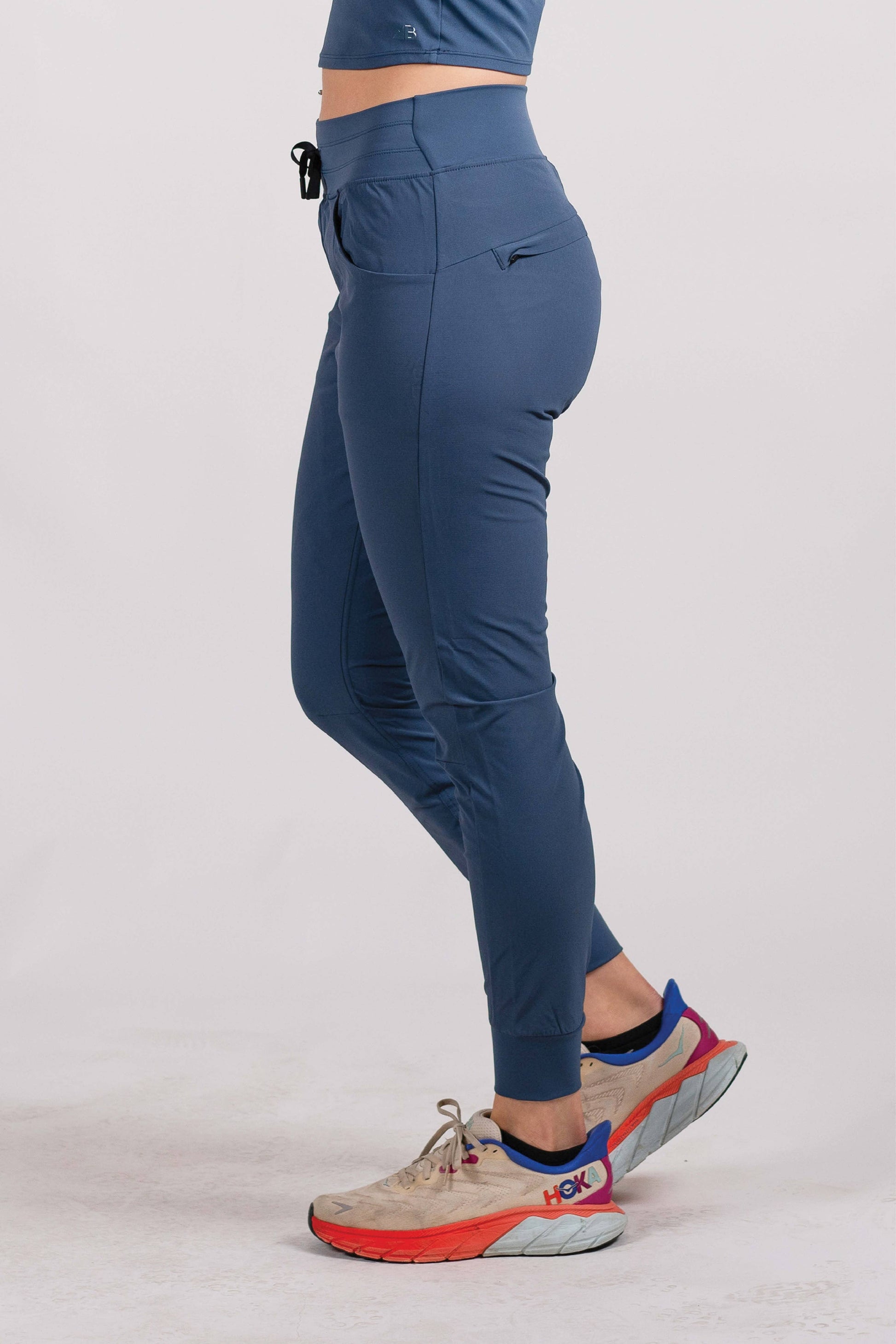 A person wearing Belong Designs' Rocky Mountain Joggers, crafted from durable and stretchy material, stands against a plain background. They are dressed in a dark green cropped top paired with matching athletic pants featuring a drawstring, complemented by brown hiking boots with teal laces. The image is cropped to show the midriff down.
