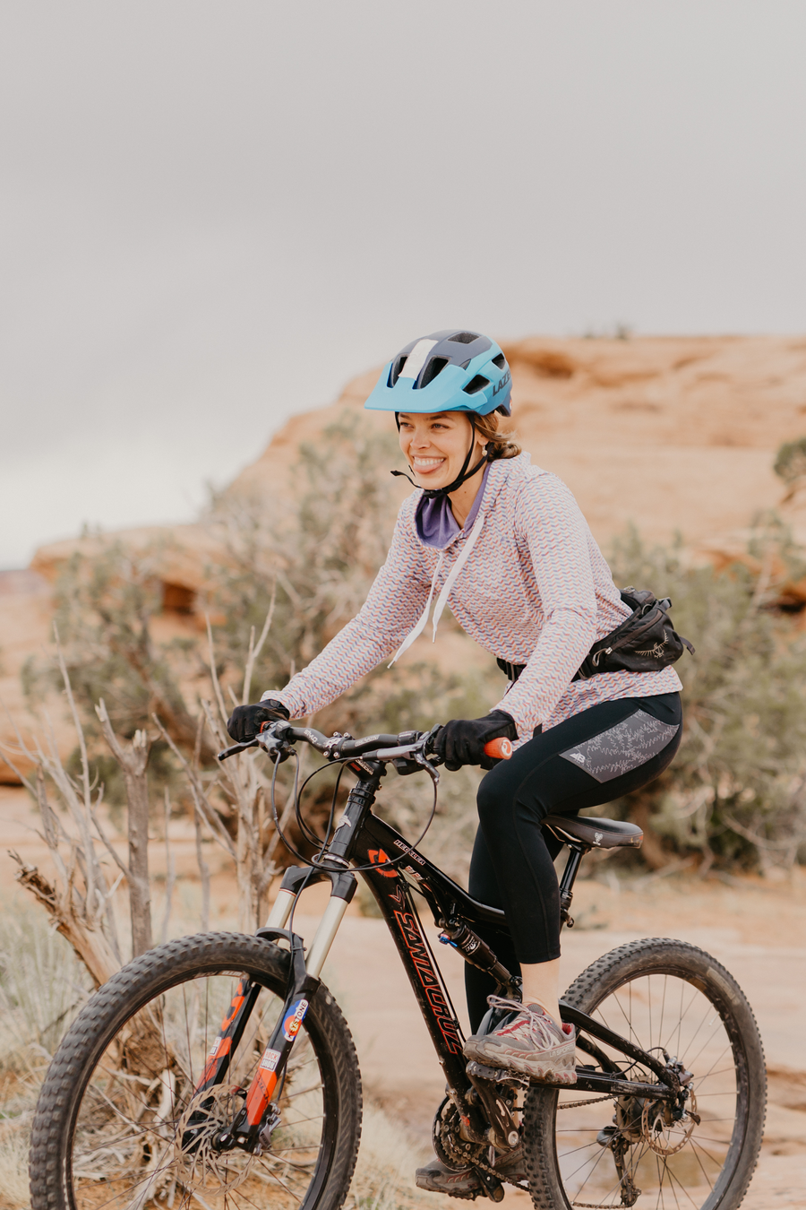 The Trailhead 7/8 Leggings from Belong Designs are black athletic leggings with a high waist and calf-length cut. They feature a colorful striped pattern in shades of purple and blue on the waist and leg panels. Crafted from PrimaFlex material, these multi-pocketed leggings also display a small logo on the left leg against a white background.