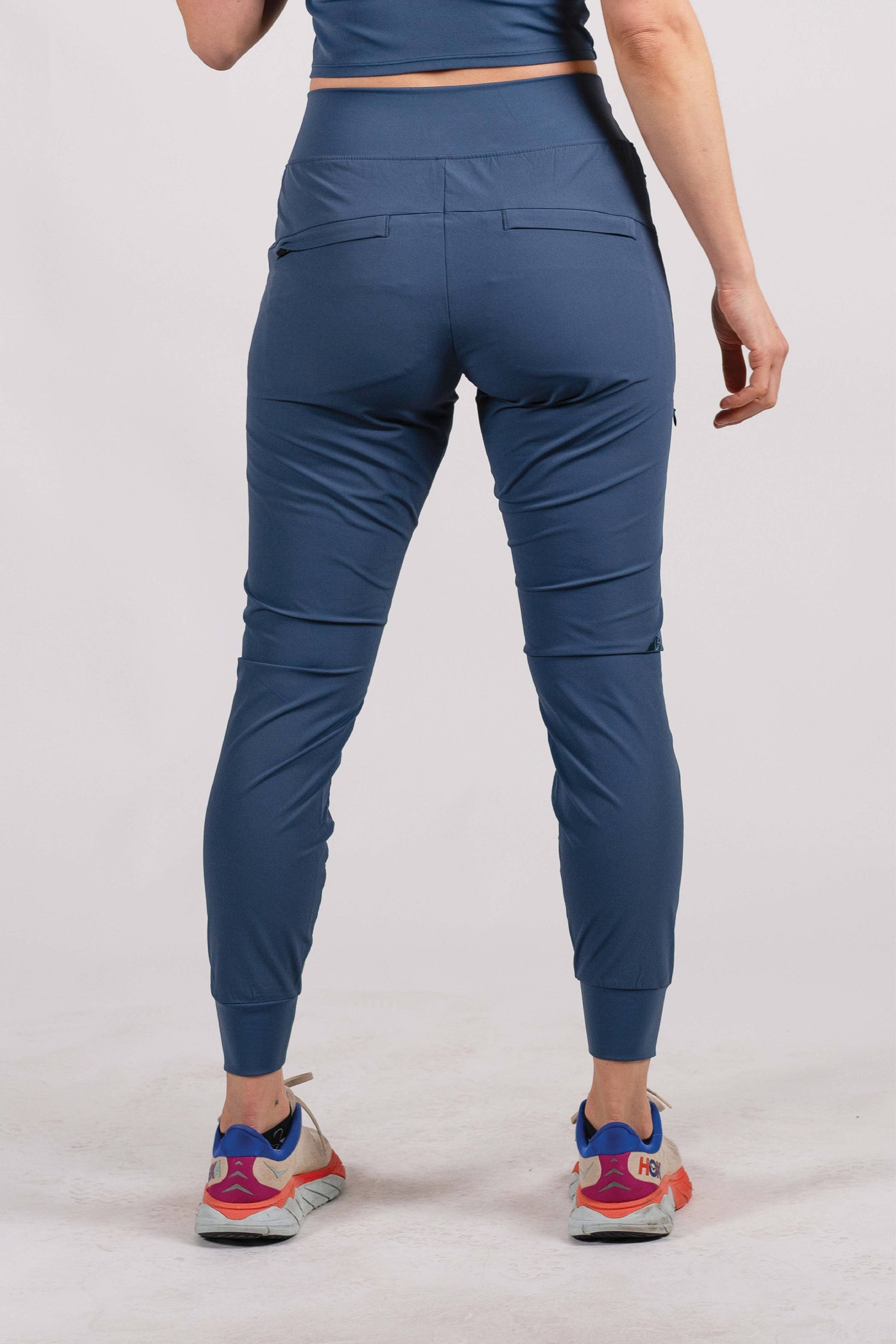 A person wearing Belong Designs' Rocky Mountain Joggers, crafted from durable and stretchy material, stands against a plain background. They are dressed in a dark green cropped top paired with matching athletic pants featuring a drawstring, complemented by brown hiking boots with teal laces. The image is cropped to show the midriff down.
