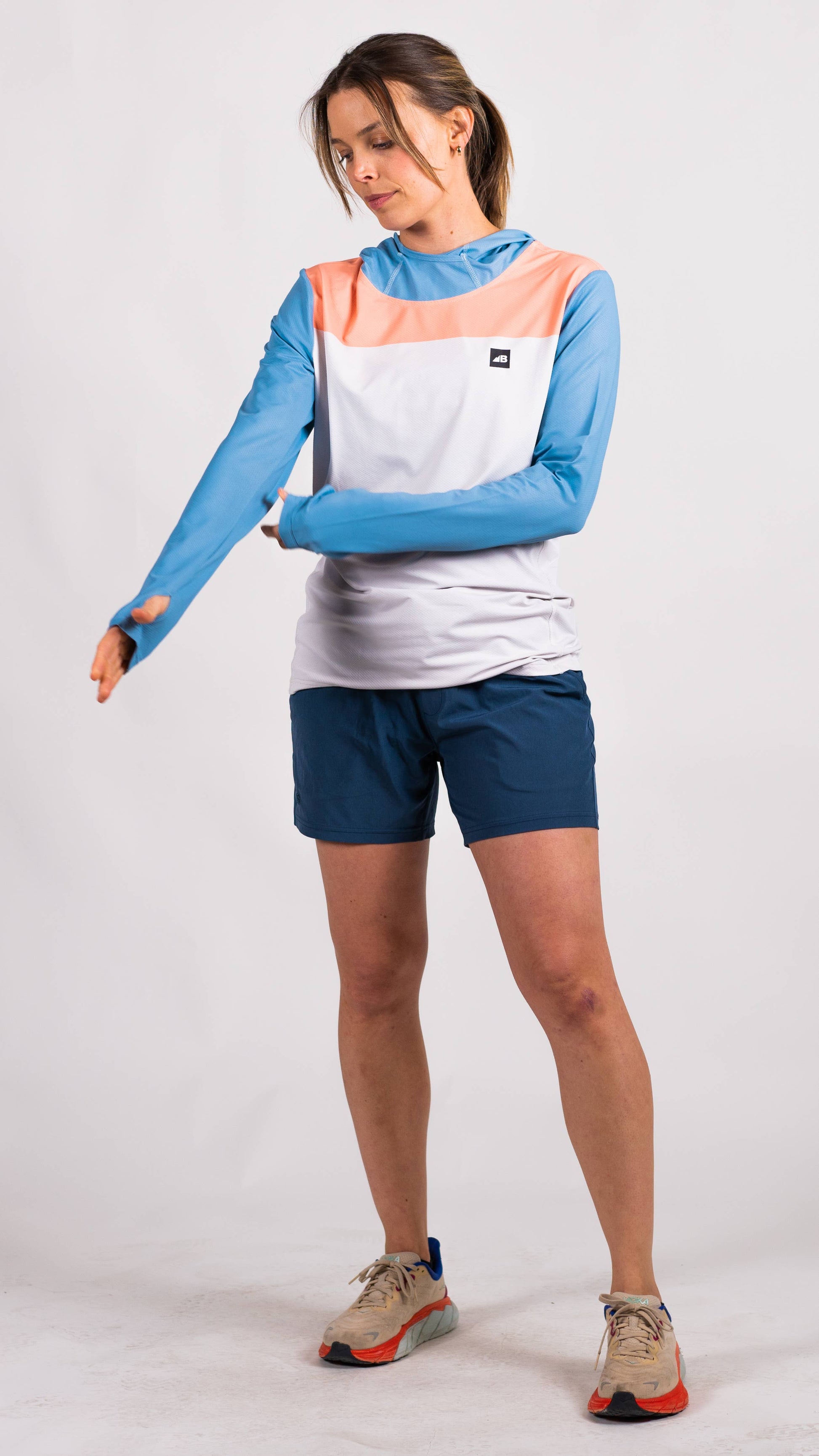 A person in a long-sleeve Sun Hoodie by Belong Designs, featuring a color-blocked design with white, maroon, and gray sections, a black logo on the chest, and wearing dark green shorts stands with hands in pockets. The background is plain white.