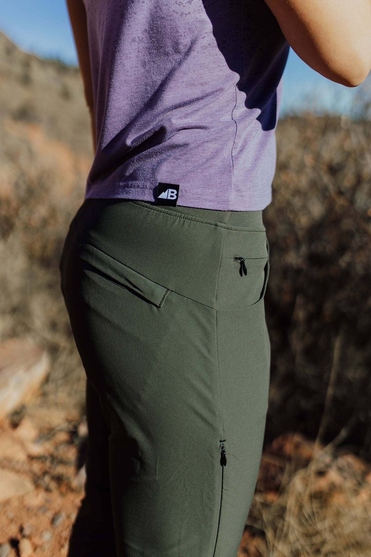 A person wearing Belong Designs' Rocky Mountain Joggers, crafted from durable and stretchy material, stands against a plain background. They are dressed in a dark green cropped top paired with matching athletic pants featuring a drawstring, complemented by brown hiking boots with teal laces. The image is cropped to show the midriff down.
