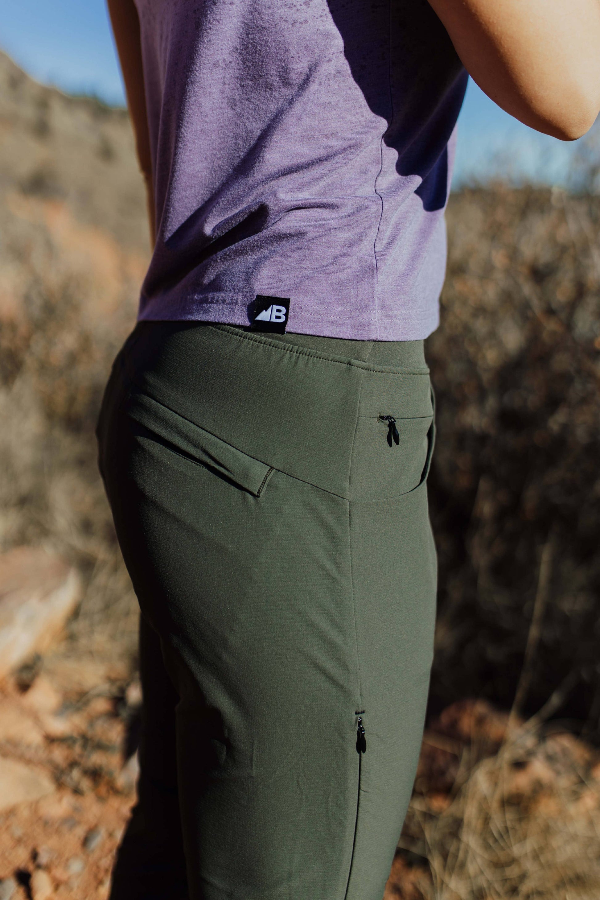 A person wearing Belong Designs' Rocky Mountain Joggers, crafted from durable and stretchy material, stands against a plain background. They are dressed in a dark green cropped top paired with matching athletic pants featuring a drawstring, complemented by brown hiking boots with teal laces. The image is cropped to show the midriff down.
