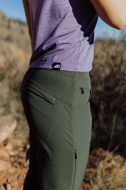 A person wearing Belong Designs' Rocky Mountain Joggers, crafted from durable and stretchy material, stands against a plain background. They are dressed in a dark green cropped top paired with matching athletic pants featuring a drawstring, complemented by brown hiking boots with teal laces. The image is cropped to show the midriff down.
