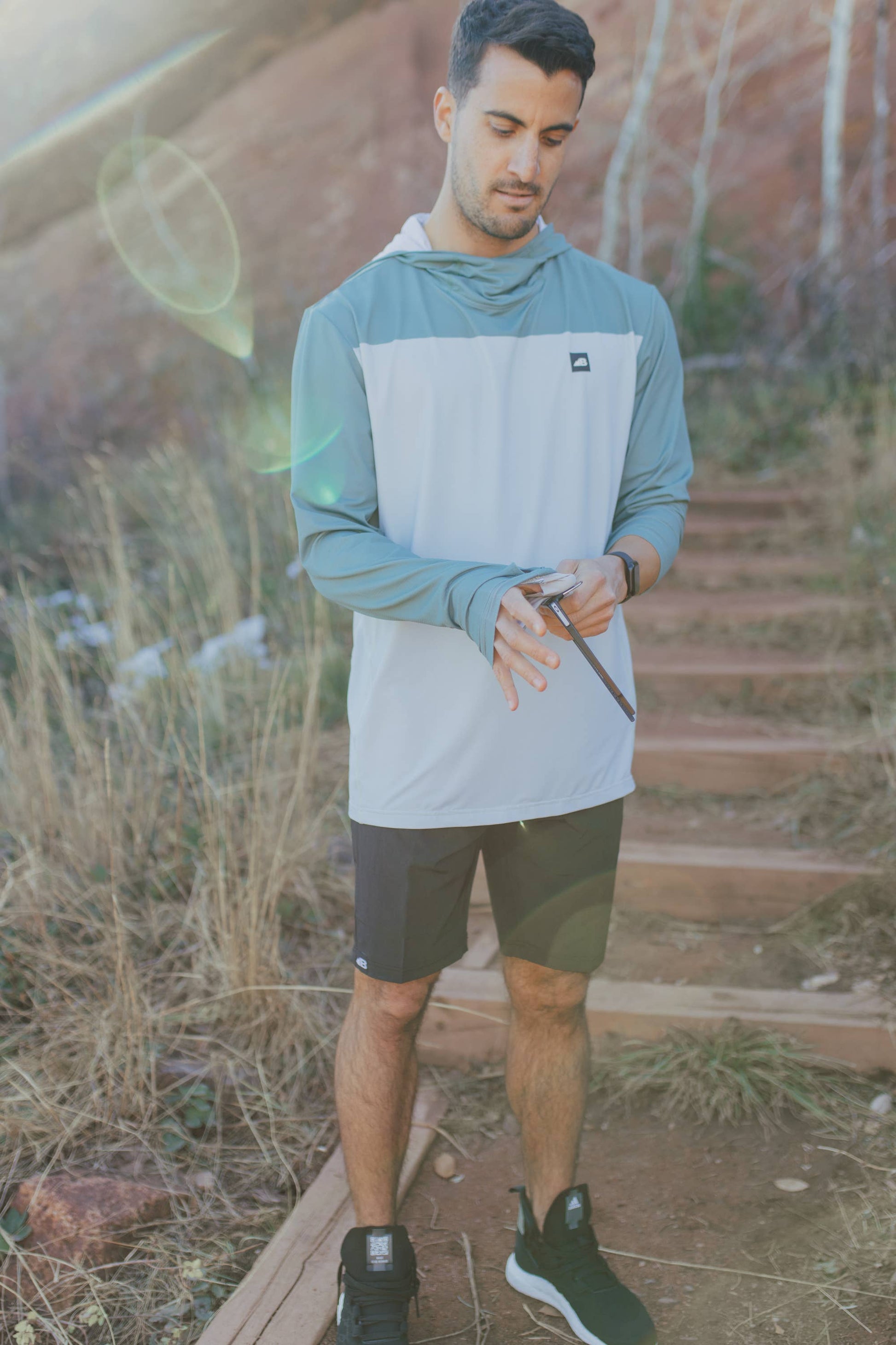 A man is wearing a Belong Designs Men's Sun Hoodie in Green Block, paired with black shorts. The hoodie features a small black logo on the left chest and offers UPF 50+ protection. He is standing against a plain, light-colored background, ready for outdoor activities. His head is not visible in the image.