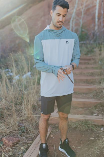A man is wearing a Belong Designs Men's Sun Hoodie in Green Block, paired with black shorts. The hoodie features a small black logo on the left chest and offers UPF 50+ protection. He is standing against a plain, light-colored background, ready for outdoor activities. His head is not visible in the image.