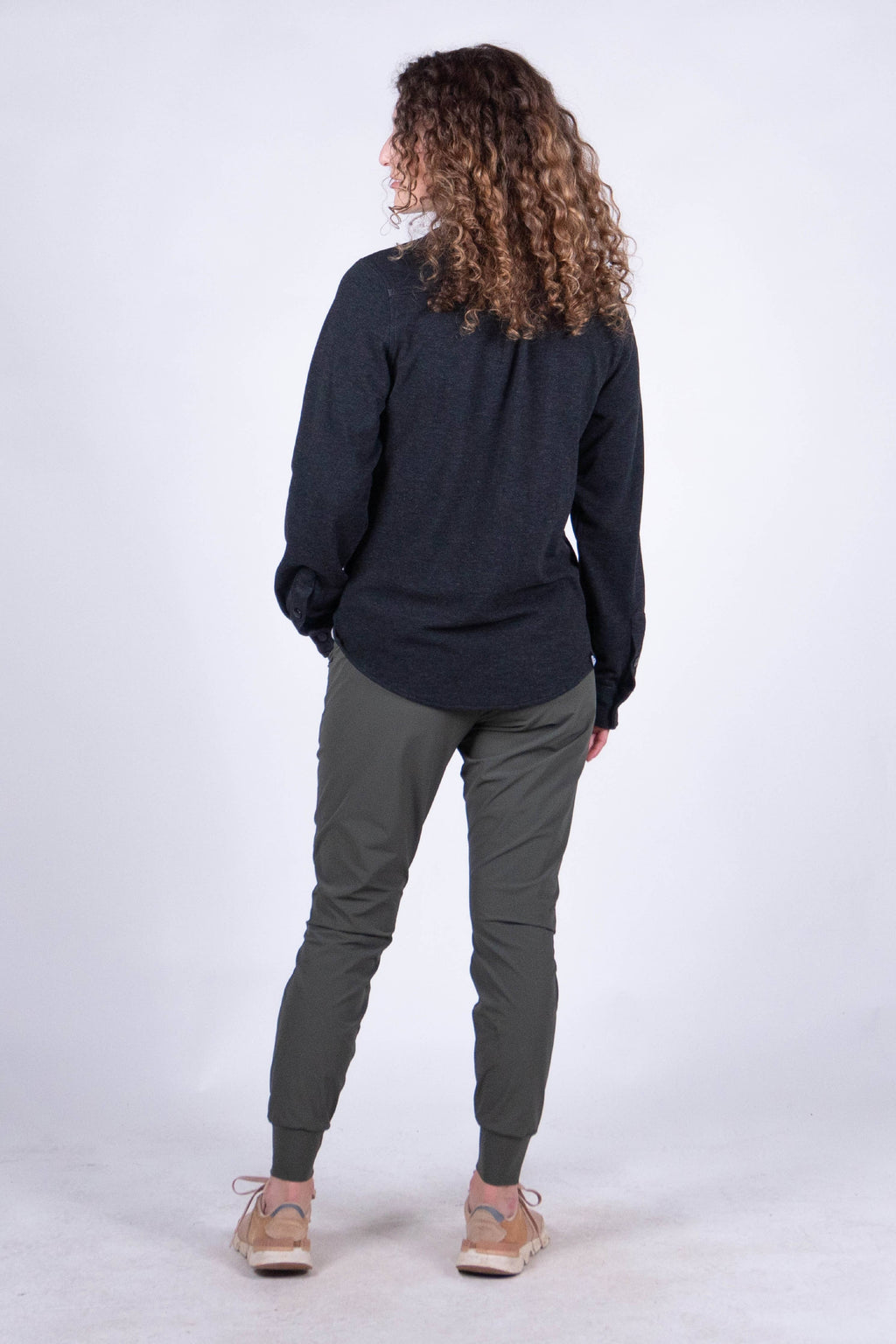 A person with curly hair stands against a plain background, smiling and looking at the camera. They are wearing the Sherman Fleece Button Up from Belong Designs, olive green pants, and beige shoes, with their hands in their pockets.