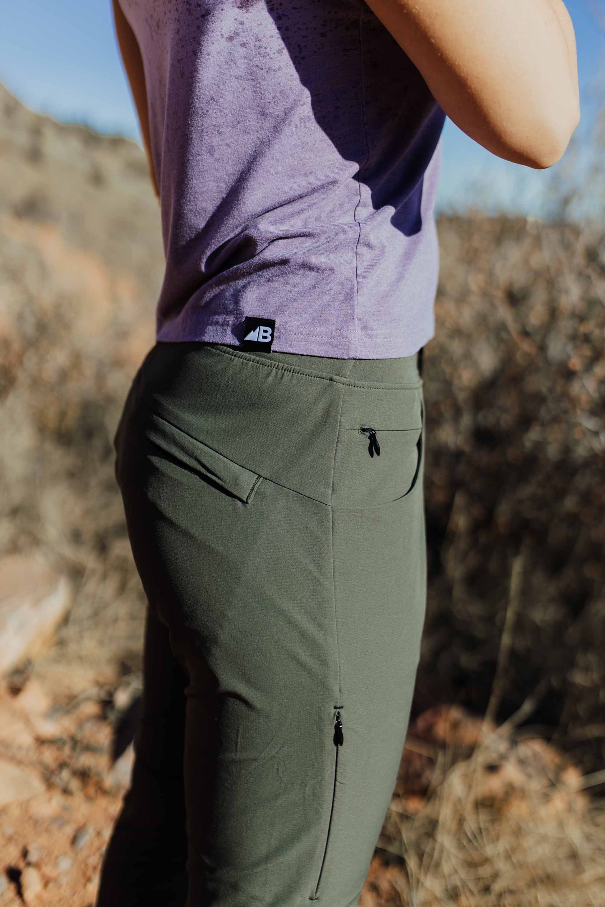 A person wearing Belong Designs' Rocky Mountain Joggers, crafted from durable and stretchy material, stands against a plain background. They are dressed in a dark green cropped top paired with matching athletic pants featuring a drawstring, complemented by brown hiking boots with teal laces. The image is cropped to show the midriff down.

