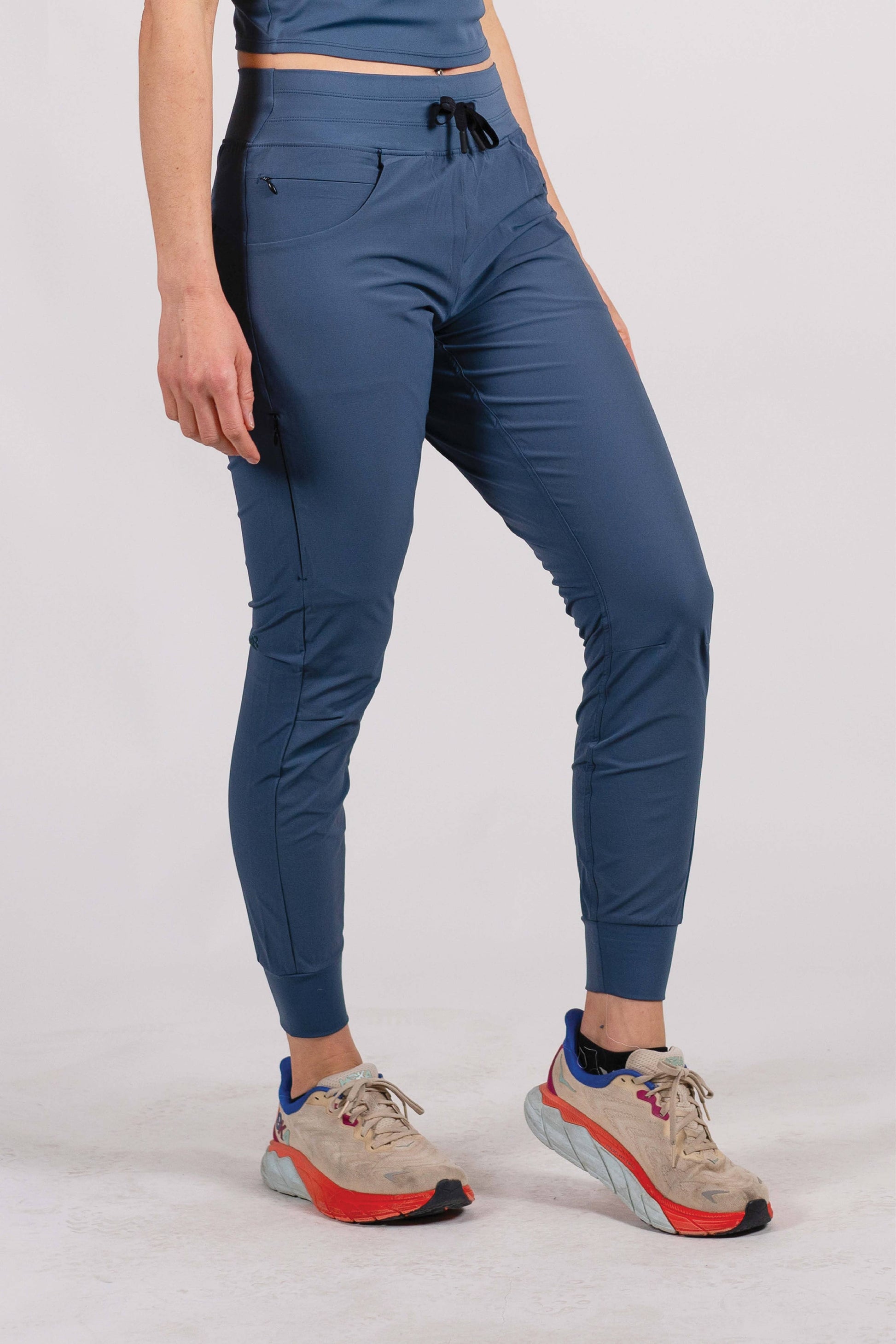 A person wearing Belong Designs' Rocky Mountain Joggers, crafted from durable and stretchy material, stands against a plain background. They are dressed in a dark green cropped top paired with matching athletic pants featuring a drawstring, complemented by brown hiking boots with teal laces. The image is cropped to show the midriff down.
