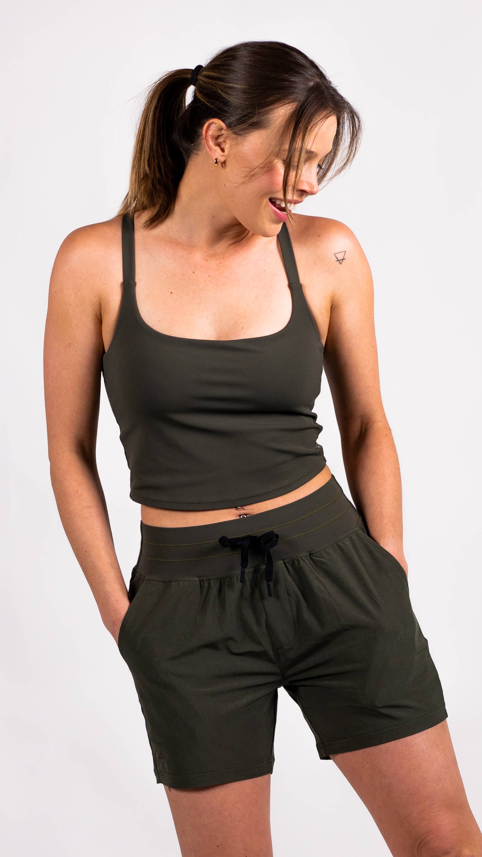 A person with long hair is dressed in a blue workout outfit from Belong Designs, consisting of the Trailhead Tek Tank in Steel Blue with adjustable straps and jogger-style pants. They are posing against a simple, white background, wearing sneakers. The person is looking to the side with one hand lightly resting near their neck.