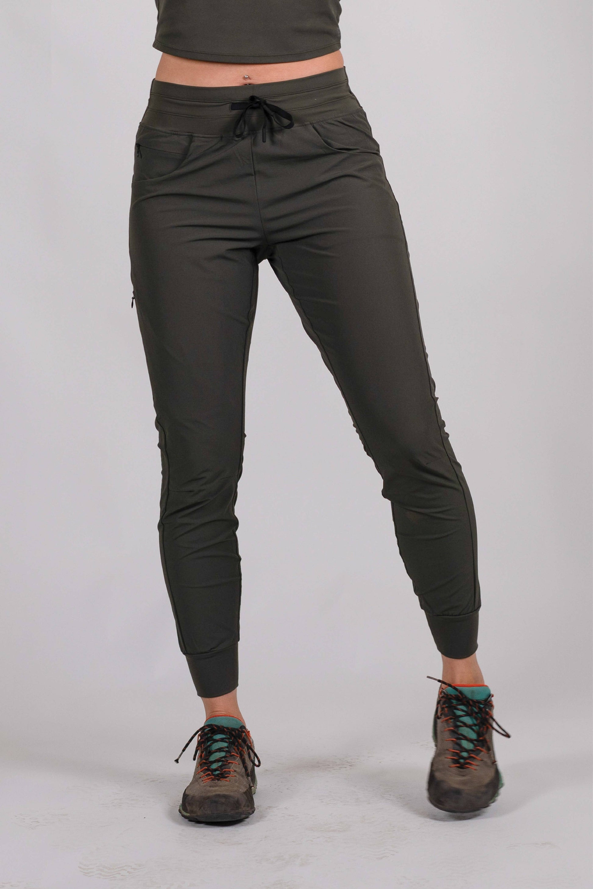 A person wearing Belong Designs' Rocky Mountain Joggers, crafted from durable and stretchy material, stands against a plain background. They are dressed in a dark green cropped top paired with matching athletic pants featuring a drawstring, complemented by brown hiking boots with teal laces. The image is cropped to show the midriff down.
