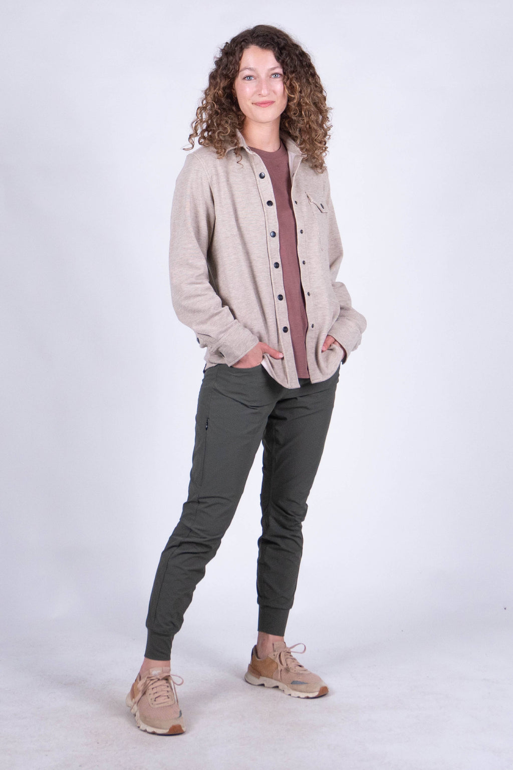 A person with curly hair stands against a plain background, smiling and looking at the camera. They are wearing the Sherman Fleece Button Up from Belong Designs, olive green pants, and beige shoes, with their hands in their pockets.