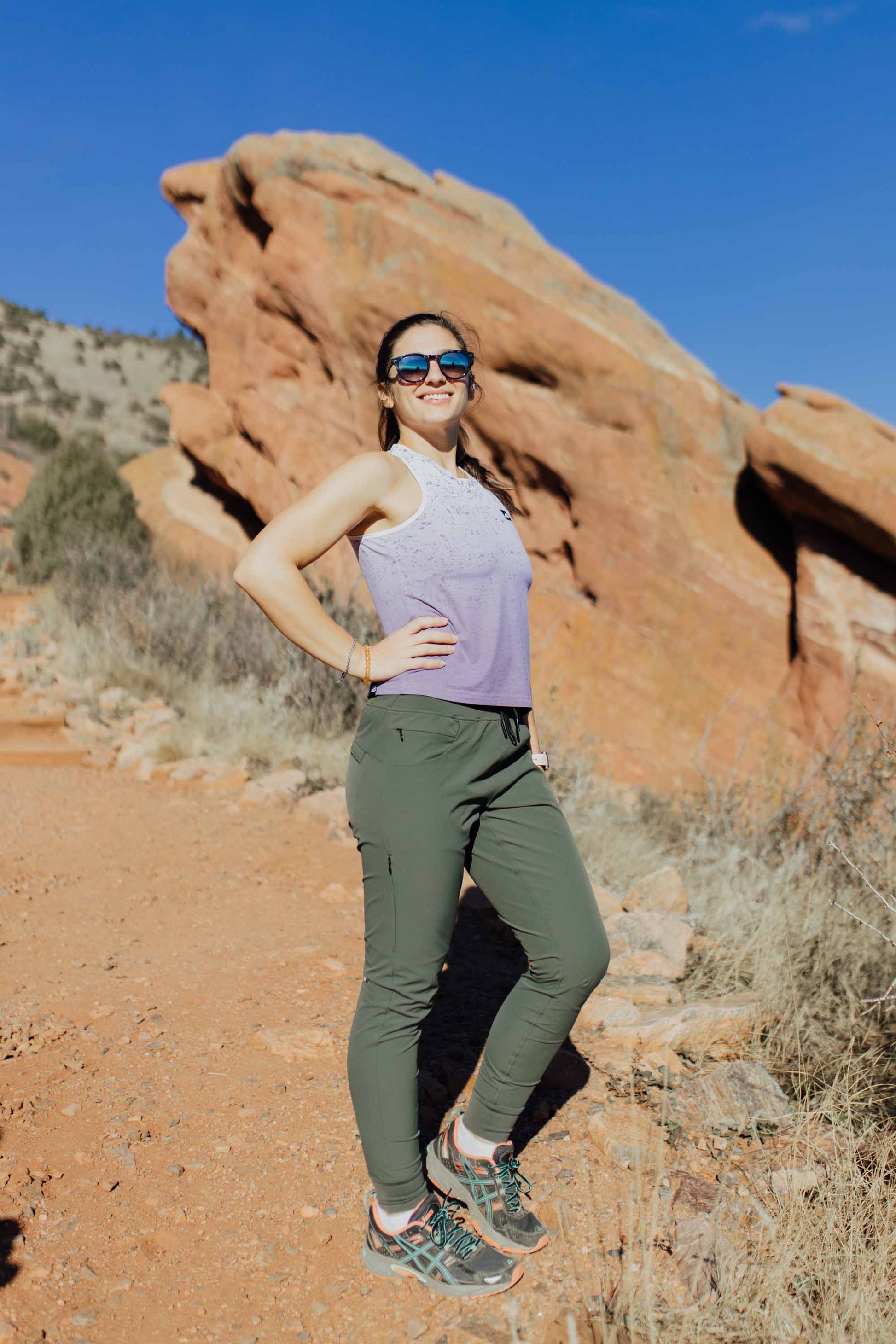 A person wearing Belong Designs' Rocky Mountain Joggers, crafted from durable and stretchy material, stands against a plain background. They are dressed in a dark green cropped top paired with matching athletic pants featuring a drawstring, complemented by brown hiking boots with teal laces. The image is cropped to show the midriff down.
