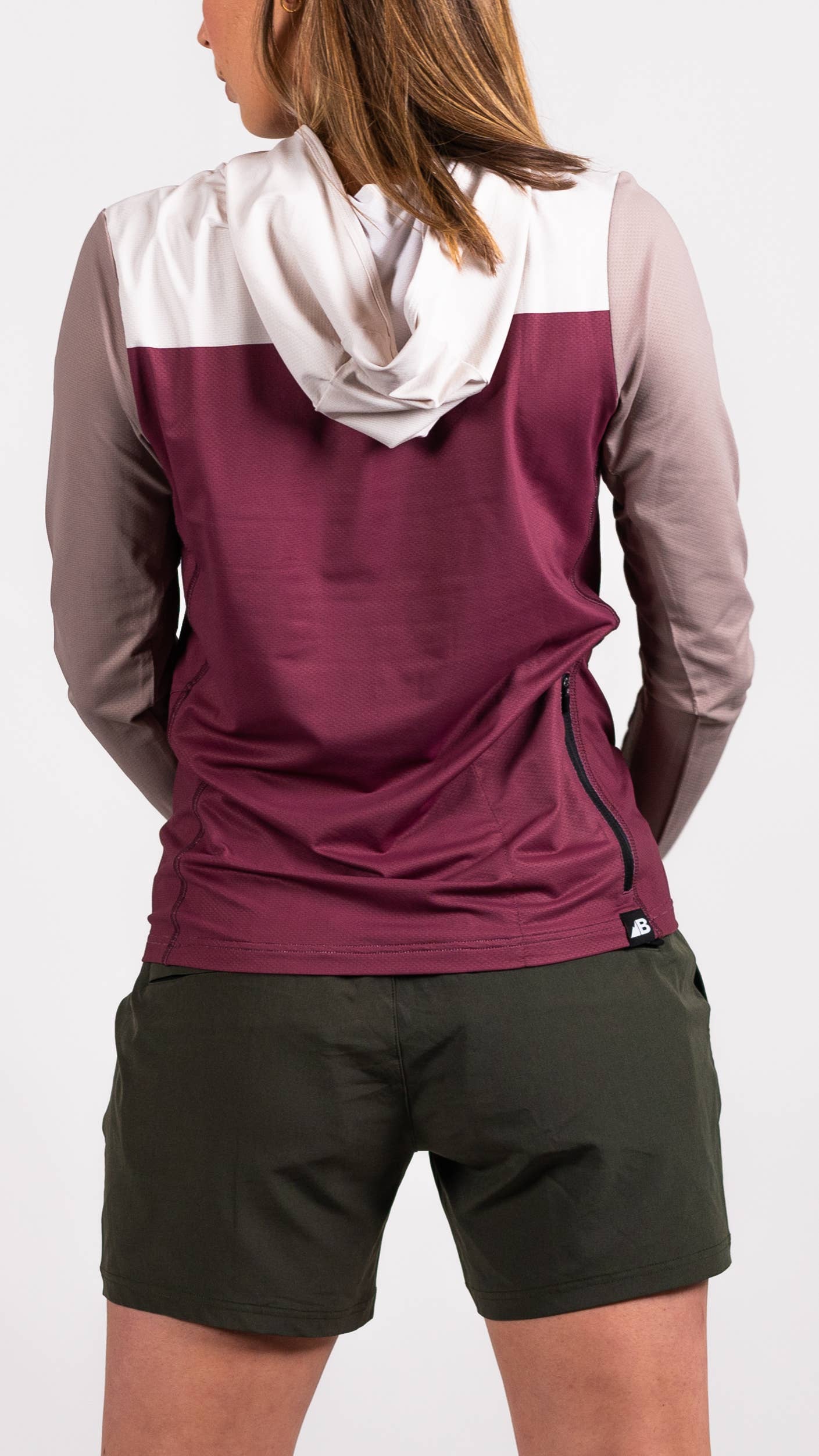 A person in a long-sleeve Sun Hoodie by Belong Designs, featuring a color-blocked design with white, maroon, and gray sections, a black logo on the chest, and wearing dark green shorts stands with hands in pockets. The background is plain white.