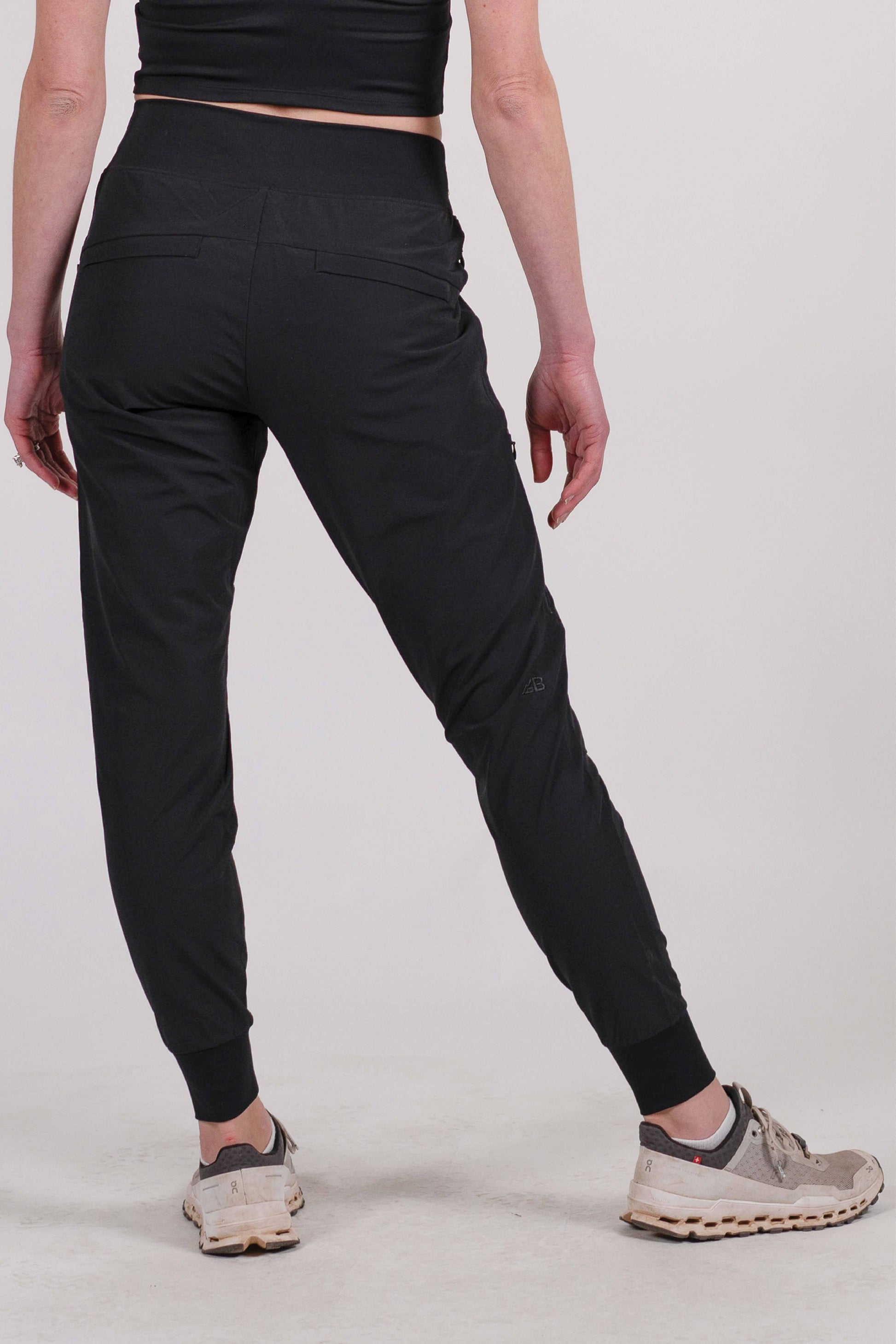 A person wearing Belong Designs' Rocky Mountain Joggers, crafted from durable and stretchy material, stands against a plain background. They are dressed in a dark green cropped top paired with matching athletic pants featuring a drawstring, complemented by brown hiking boots with teal laces. The image is cropped to show the midriff down.

