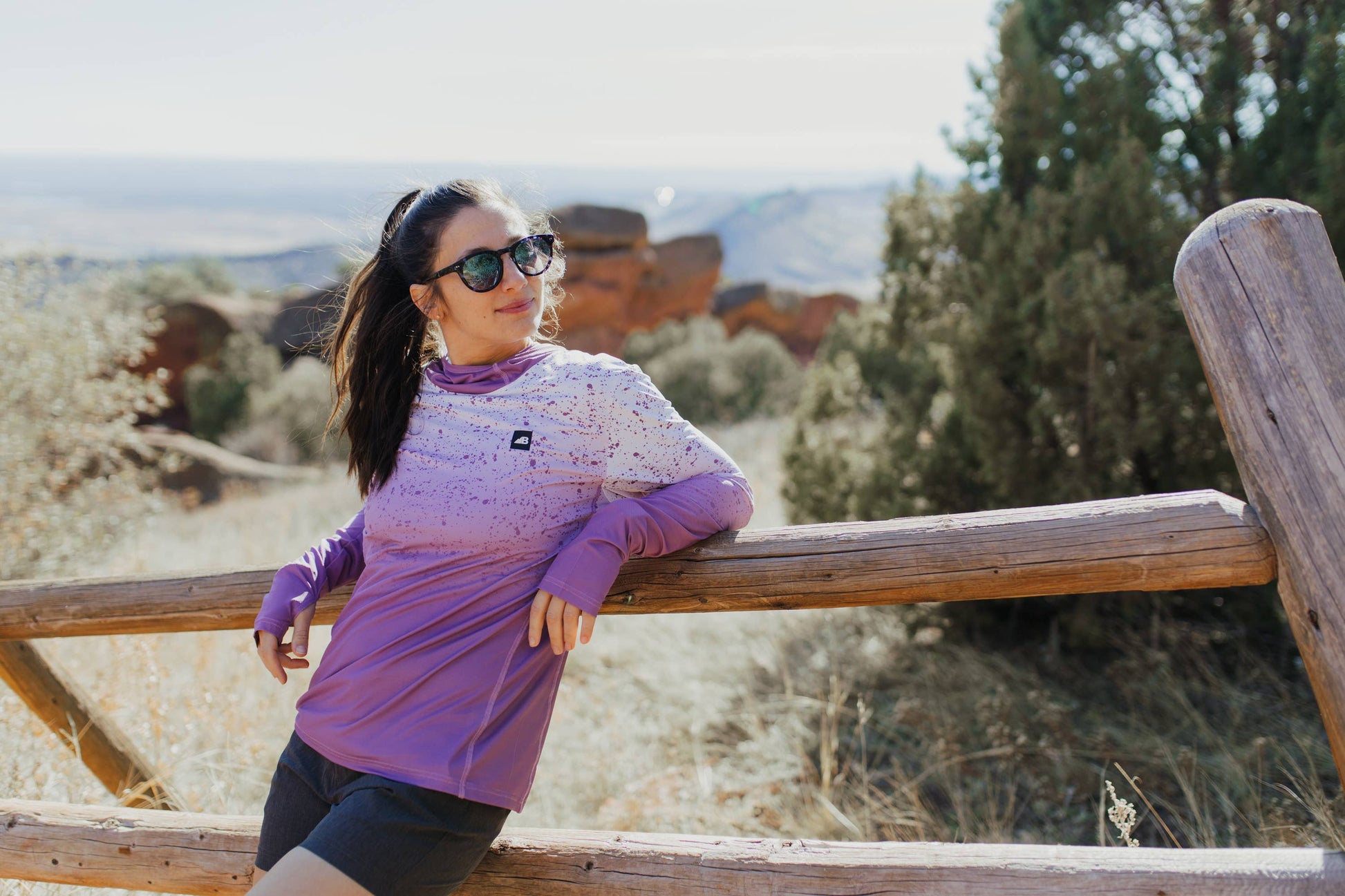A person in a long-sleeve Sun Hoodie by Belong Designs, featuring a color-blocked design with white, maroon, and gray sections, a black logo on the chest, and wearing dark green shorts stands with hands in pockets. The background is plain white.
