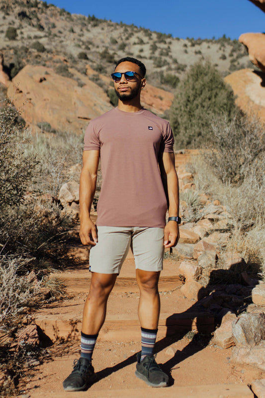 A man stands on a rocky trail with mountainous terrain in the background, wearing the mauve Men's Core Traverse Tek Tee from Belong Designs, light gray athletic fit shorts, dark socks, black shoes, and reflective sunglasses. The sky is clear and blue, with dry shrubs surrounding the trail.