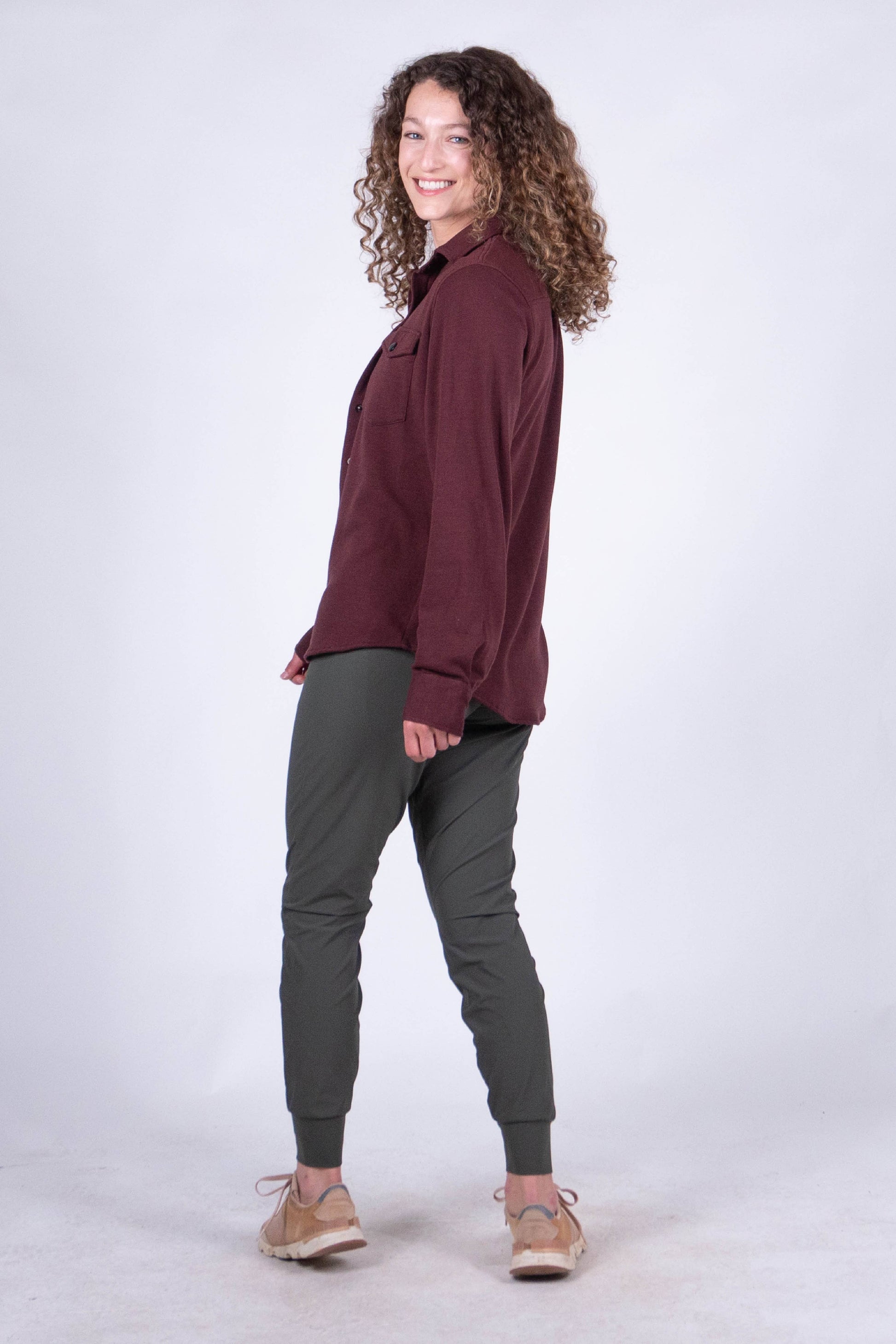 A person with curly hair stands against a plain background, smiling and looking at the camera. They are wearing the Sherman Fleece Button Up from Belong Designs, olive green pants, and beige shoes, with their hands in their pockets.