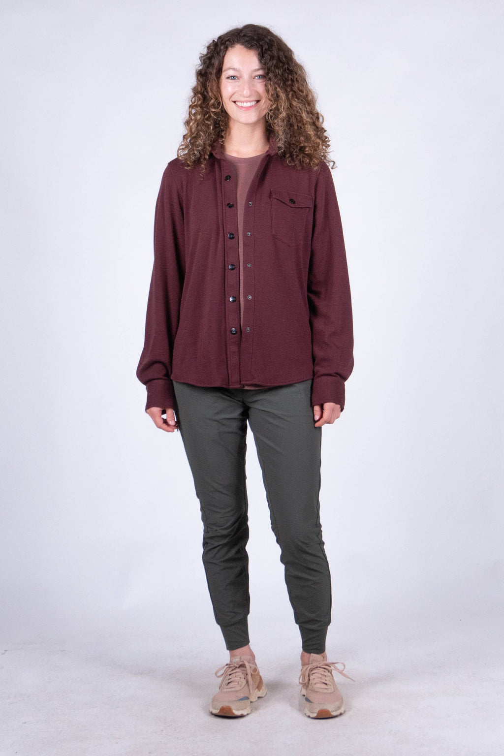 A person with curly hair stands against a plain background, smiling and looking at the camera. They are wearing the Sherman Fleece Button Up from Belong Designs, olive green pants, and beige shoes, with their hands in their pockets.