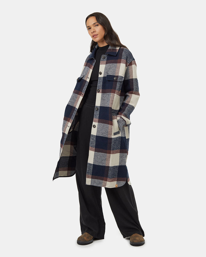 A person wearing the Tentree Heavy Weight Flannel Long Jacket made from recycled materials, featuring large blue, white, grey, and burgundy checks. With one hand in a pocket and the other resting by their side, they stand against a plain background with long hair flowing down.