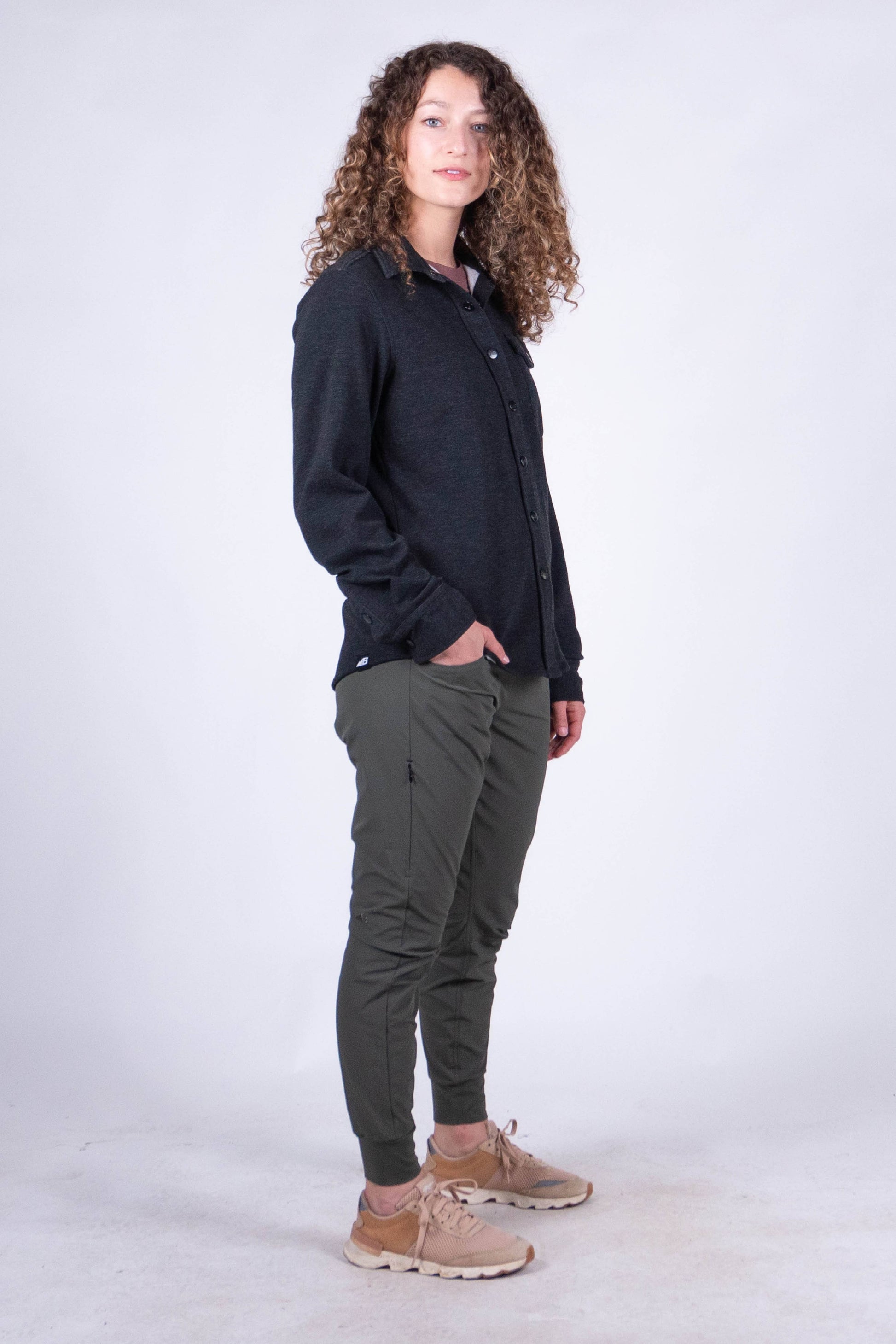 A person with curly hair stands against a plain background, smiling and looking at the camera. They are wearing the Sherman Fleece Button Up from Belong Designs, olive green pants, and beige shoes, with their hands in their pockets.