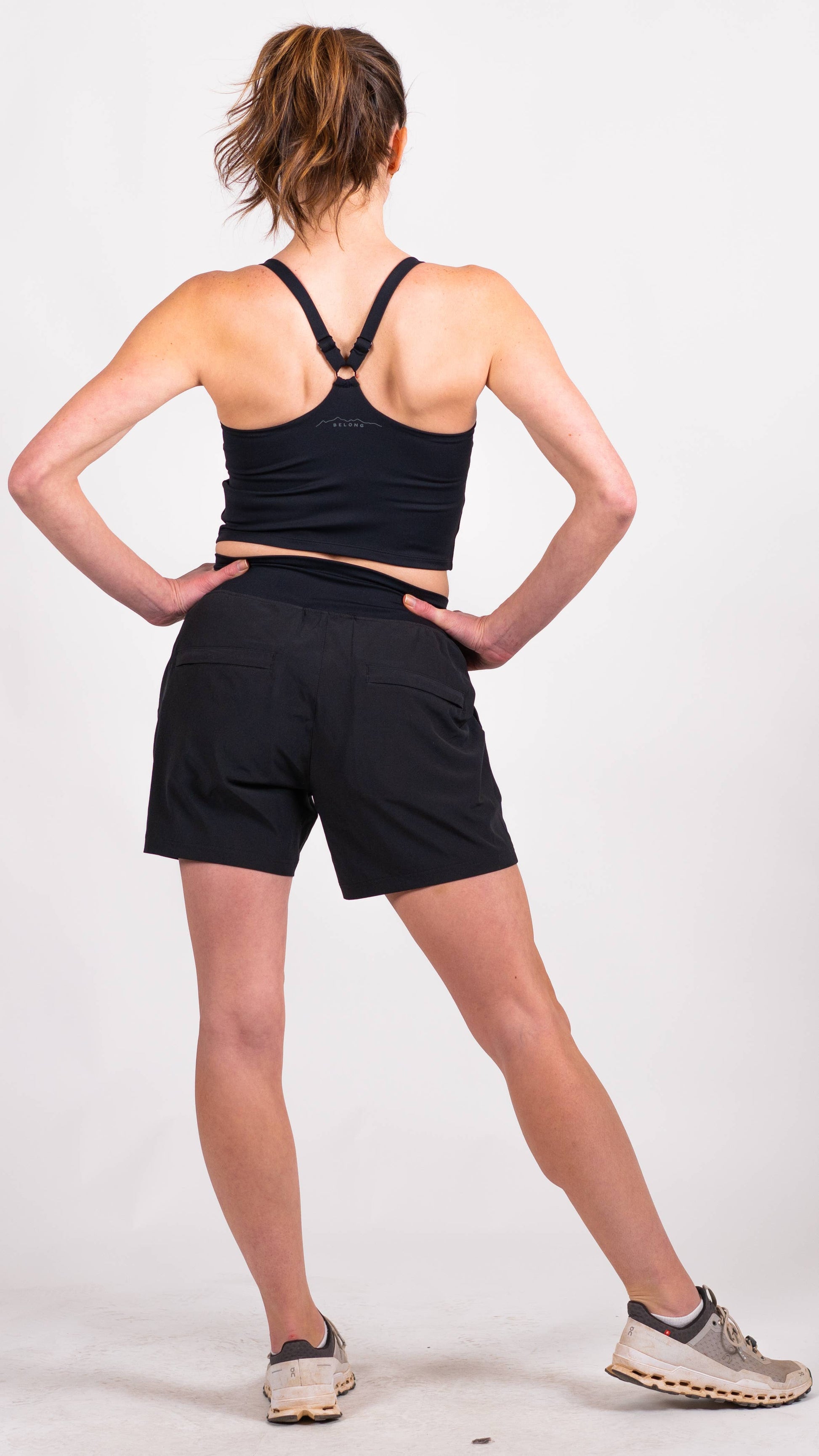 A person with long hair is dressed in a blue workout outfit from Belong Designs, consisting of the Trailhead Tek Tank in Steel Blue with adjustable straps and jogger-style pants. They are posing against a simple, white background, wearing sneakers. The person is looking to the side with one hand lightly resting near their neck.