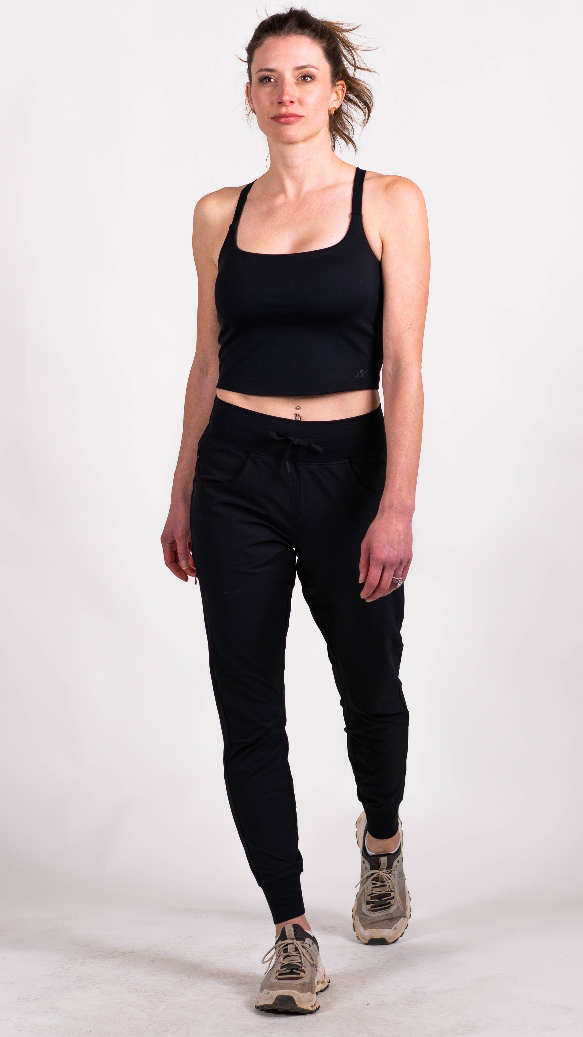 A person with long hair is dressed in a blue workout outfit from Belong Designs, consisting of the Trailhead Tek Tank in Steel Blue with adjustable straps and jogger-style pants. They are posing against a simple, white background, wearing sneakers. The person is looking to the side with one hand lightly resting near their neck.