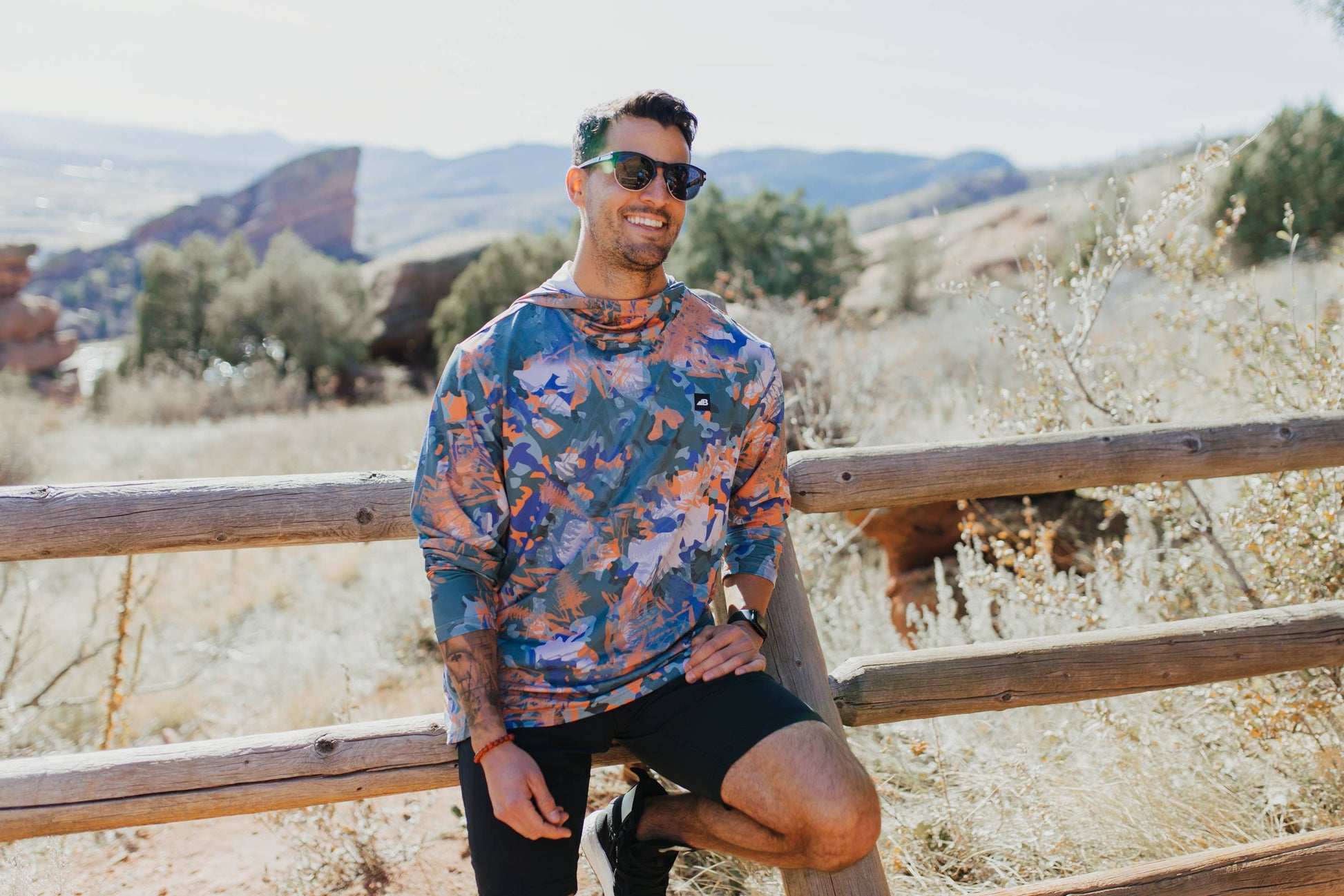 A man is wearing a Belong Designs Men's Sun Hoodie in Green Block, paired with black shorts. The hoodie features a small black logo on the left chest and offers UPF 50+ protection. He is standing against a plain, light-colored background, ready for outdoor activities. His head is not visible in the image.