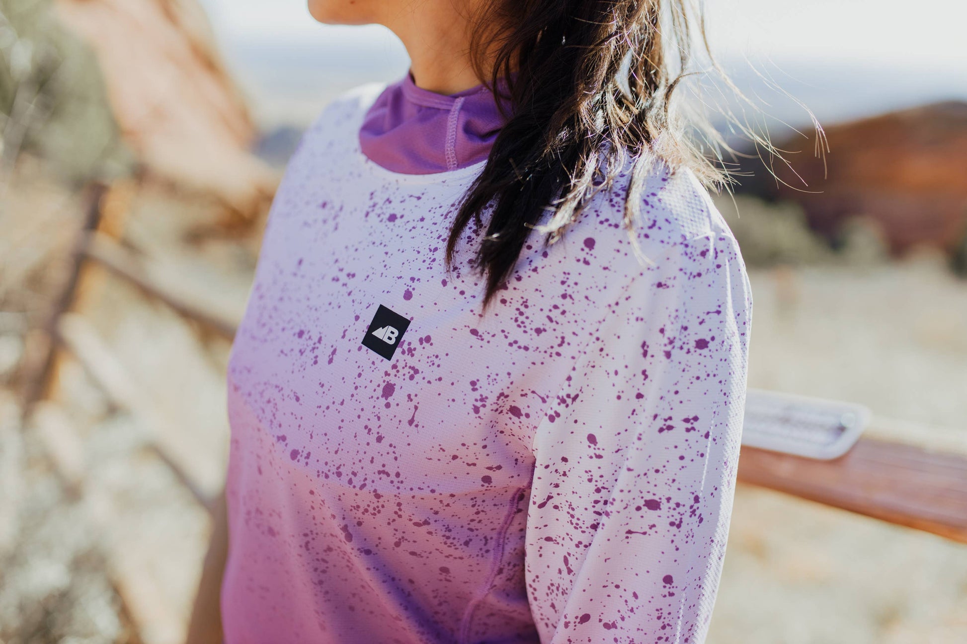 A person in a long-sleeve Sun Hoodie by Belong Designs, featuring a color-blocked design with white, maroon, and gray sections, a black logo on the chest, and wearing dark green shorts stands with hands in pockets. The background is plain white.