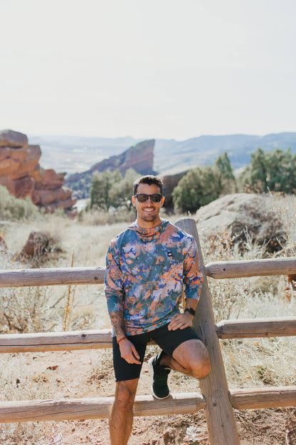 A man is wearing a Belong Designs Men's Sun Hoodie in Green Block, paired with black shorts. The hoodie features a small black logo on the left chest and offers UPF 50+ protection. He is standing against a plain, light-colored background, ready for outdoor activities. His head is not visible in the image.