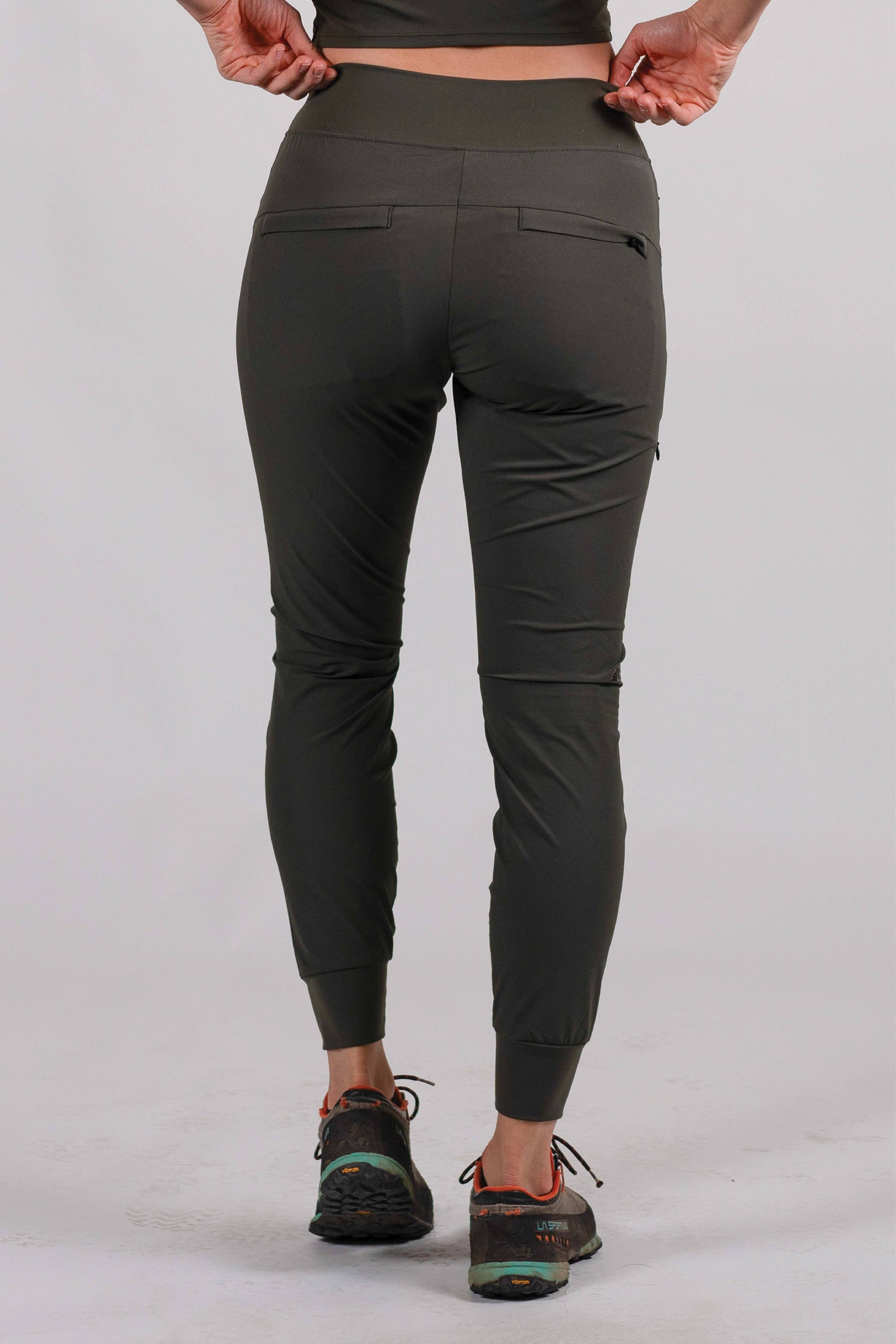 A person wearing Belong Designs' Rocky Mountain Joggers, crafted from durable and stretchy material, stands against a plain background. They are dressed in a dark green cropped top paired with matching athletic pants featuring a drawstring, complemented by brown hiking boots with teal laces. The image is cropped to show the midriff down.
