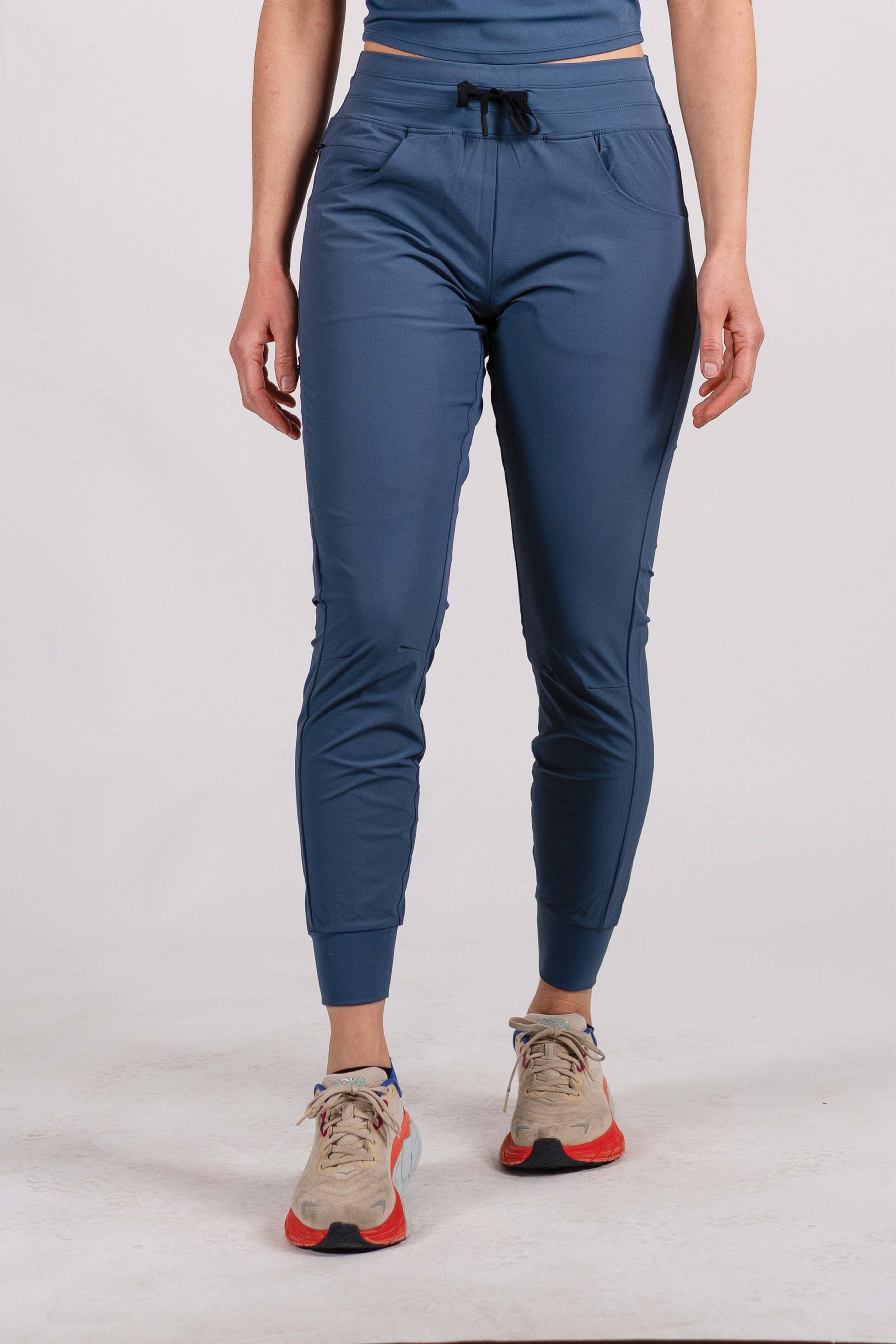 A person wearing Belong Designs' Rocky Mountain Joggers, crafted from durable and stretchy material, stands against a plain background. They are dressed in a dark green cropped top paired with matching athletic pants featuring a drawstring, complemented by brown hiking boots with teal laces. The image is cropped to show the midriff down.
