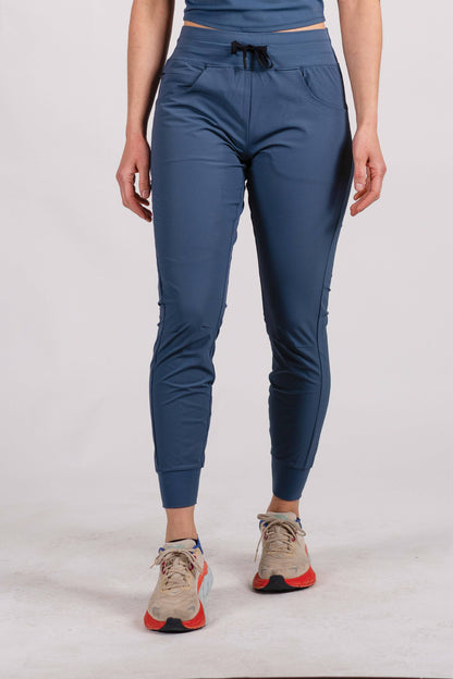 A person wearing Belong Designs' Rocky Mountain Joggers, crafted from durable and stretchy material, stands against a plain background. They are dressed in a dark green cropped top paired with matching athletic pants featuring a drawstring, complemented by brown hiking boots with teal laces. The image is cropped to show the midriff down.

