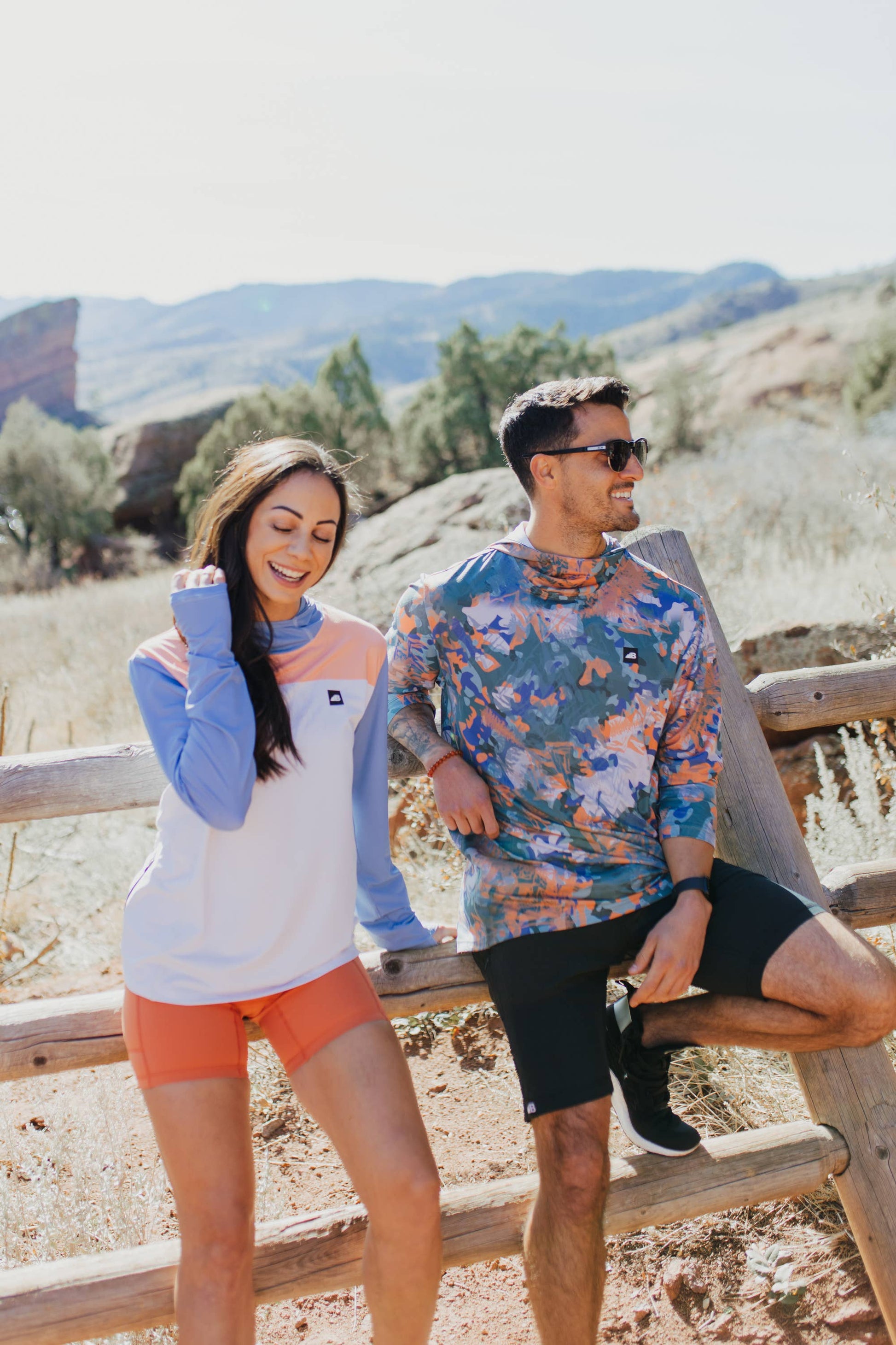 A person in a long-sleeve Sun Hoodie by Belong Designs, featuring a color-blocked design with white, maroon, and gray sections, a black logo on the chest, and wearing dark green shorts stands with hands in pockets. The background is plain white.