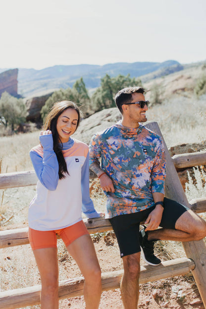 A person in a long-sleeve Sun Hoodie by Belong Designs, featuring a color-blocked design with white, maroon, and gray sections, a black logo on the chest, and wearing dark green shorts stands with hands in pockets. The background is plain white.