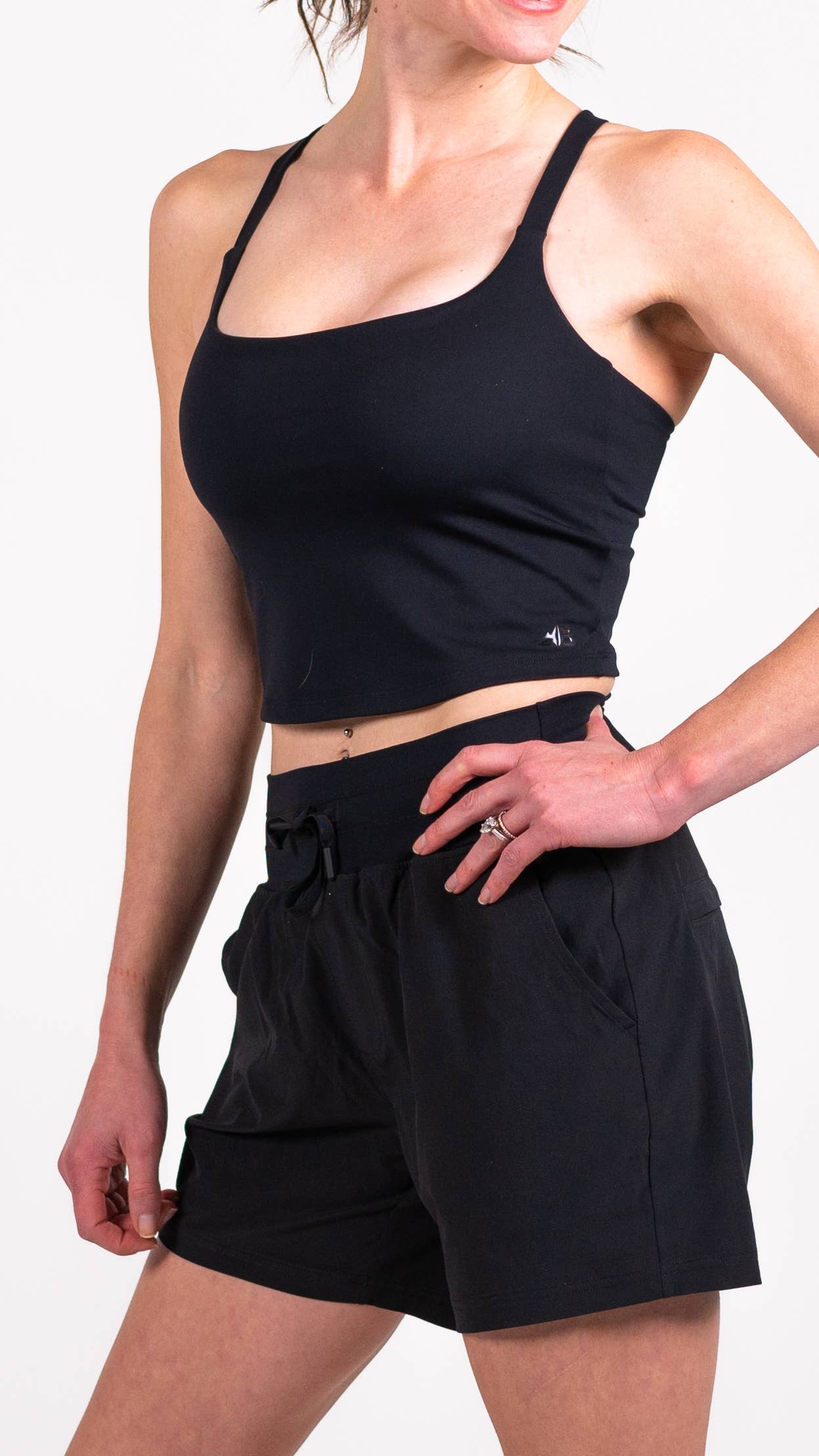 A person with long hair is dressed in a blue workout outfit from Belong Designs, consisting of the Trailhead Tek Tank in Steel Blue with adjustable straps and jogger-style pants. They are posing against a simple, white background, wearing sneakers. The person is looking to the side with one hand lightly resting near their neck.