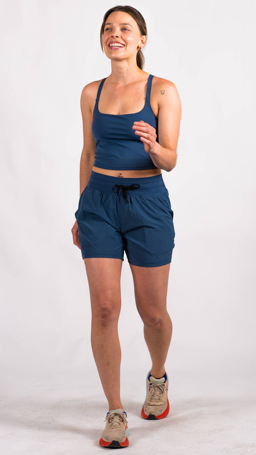 A woman dressed in blue activewear, featuring a sleeveless top and Belong Designs' 5" La Plata High Rise Hiking Shorts made with quick-drying technology, is walking. She is wearing sneakers and smiling, with one arm bent and the other relaxed at her side. The background is plain white.